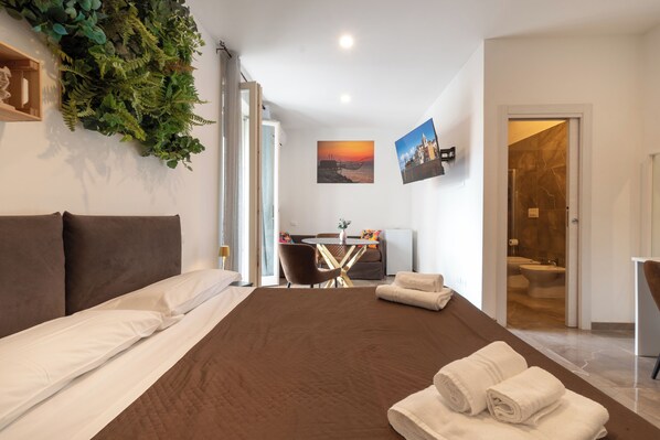 A hotel room with a bed, a desk, a chair, a bathroom, and a view of the sunset. - Suebi Central - Deluxe Rooms & Family Suites (Termoli)