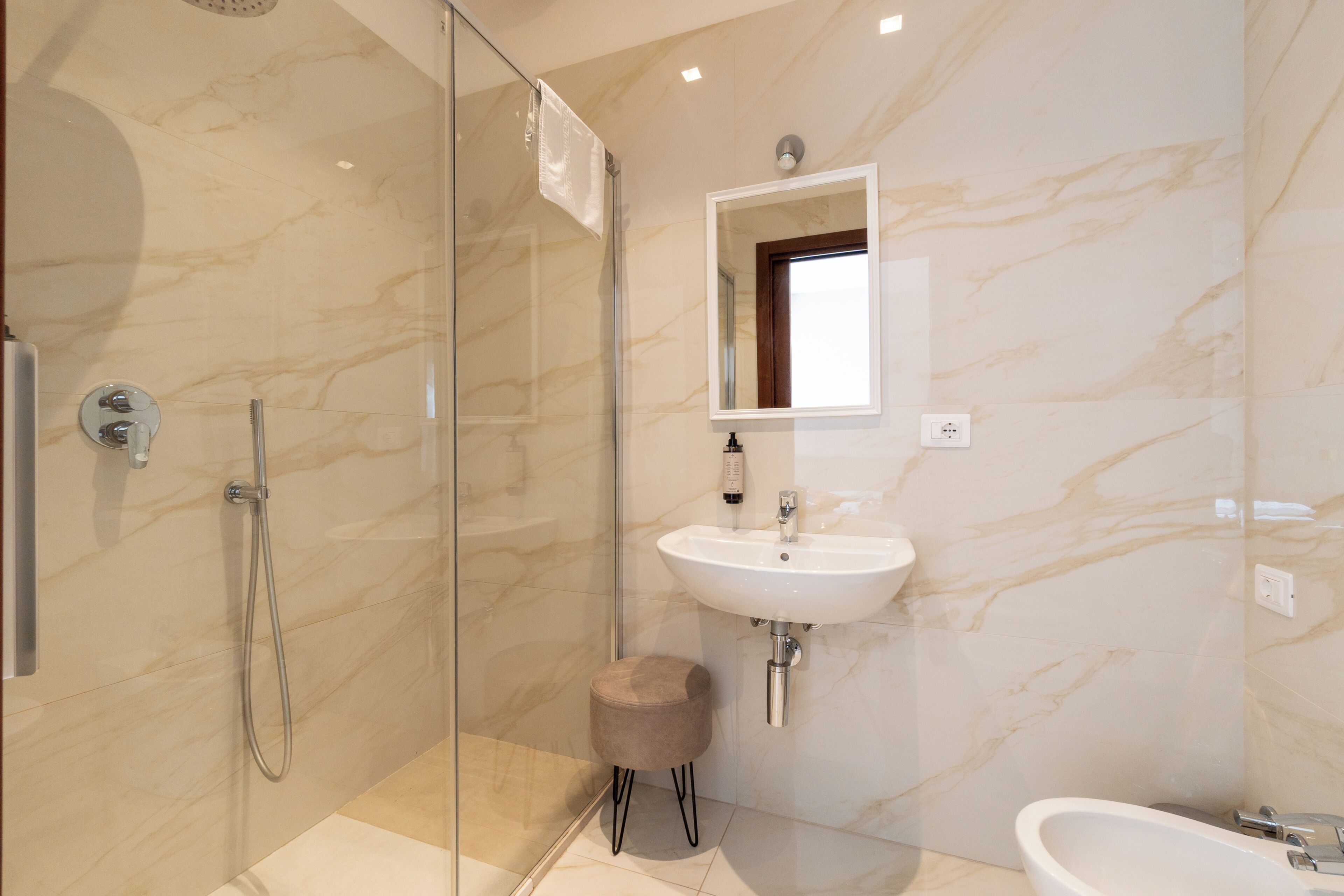 A modern bathroom with a glass shower enclosure, a pedestal sink, and a marble wall.