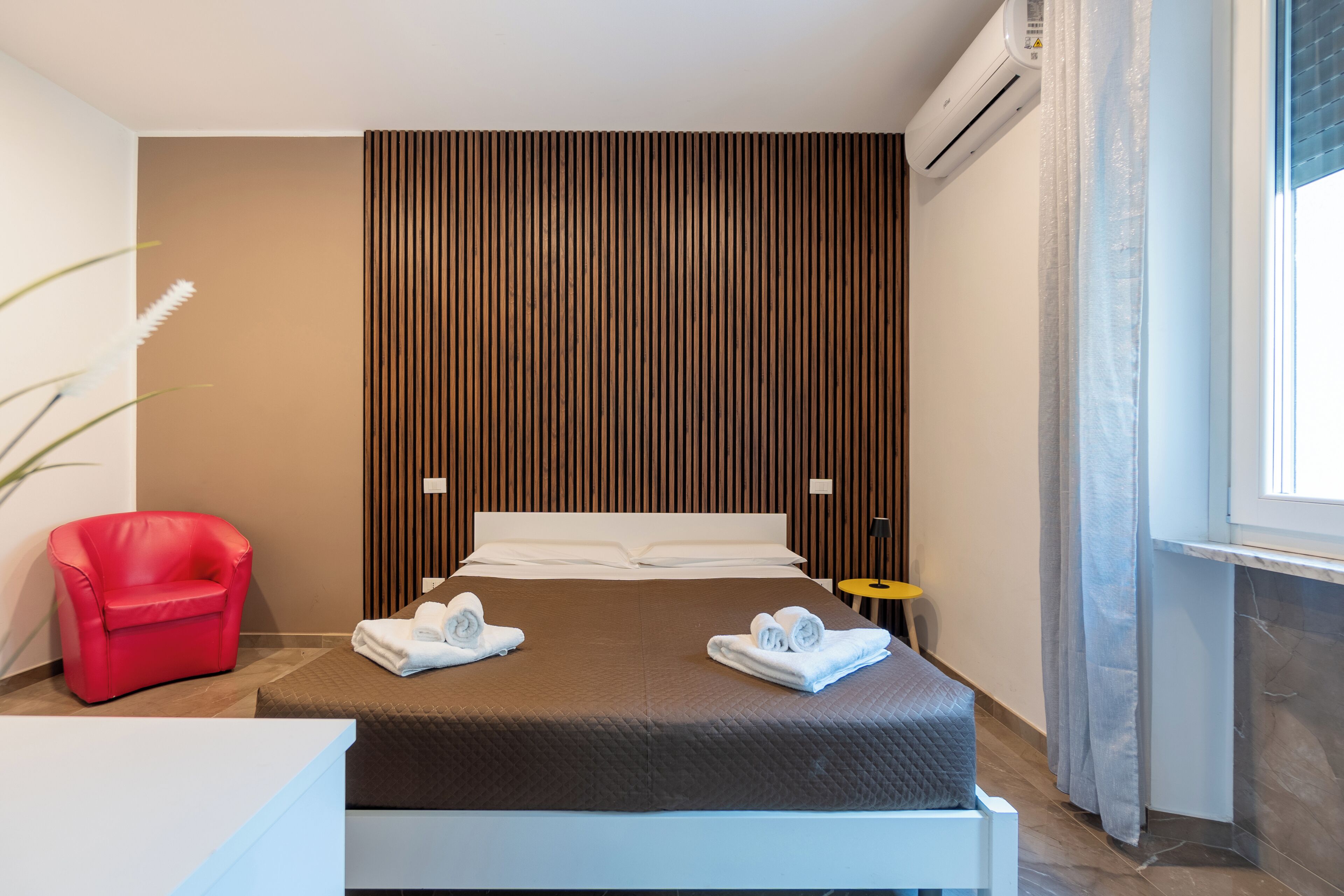A modern hotel room with a large bed, a red armchair, a small table, and a window with curtains.