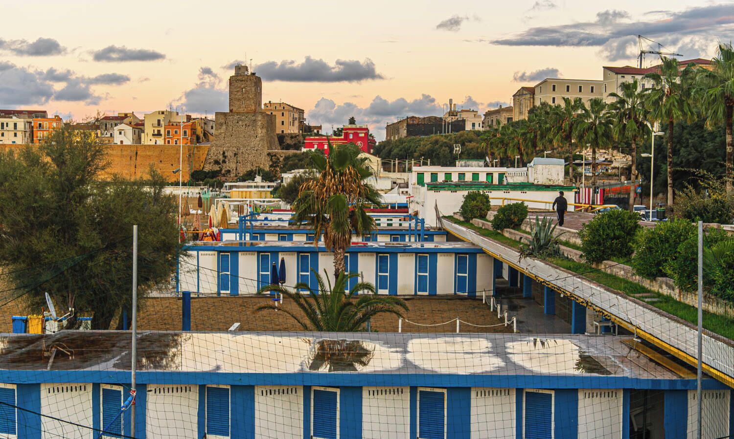 A coastal hotel with multiple blue-roofed rooms, a palm tree, and a historic tower in the background.
