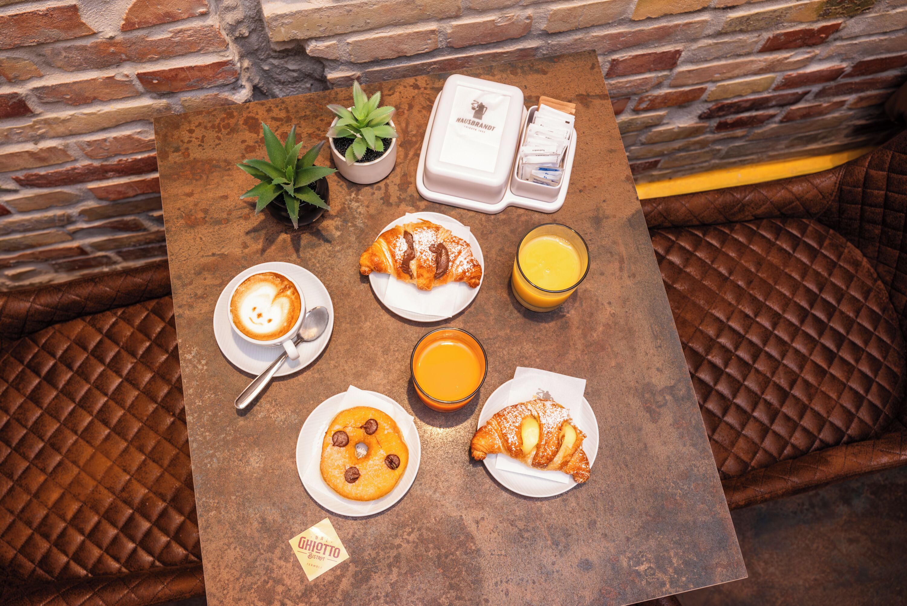 A table with breakfast items including croissants, a coffee drink, and orange juice, set against a brick wall.
