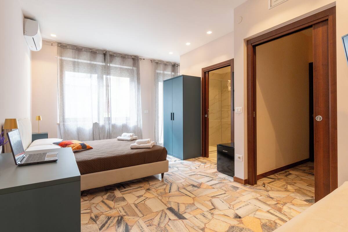 A bedroom with a bed, a desk with a laptop, a wardrobe, and a bathroom with a shower.