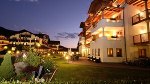 A multi-story hotel with balconies, outdoor seating, and a well-lit garden area. - Hotel Nele (Ziano di Fiemme)