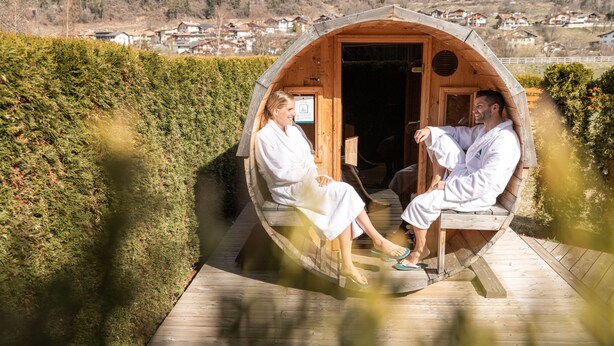A wooden sauna with two people sitting inside, surrounded by a wooden deck and a hedge. - Hotel Nele (Ziano di Fiemme)