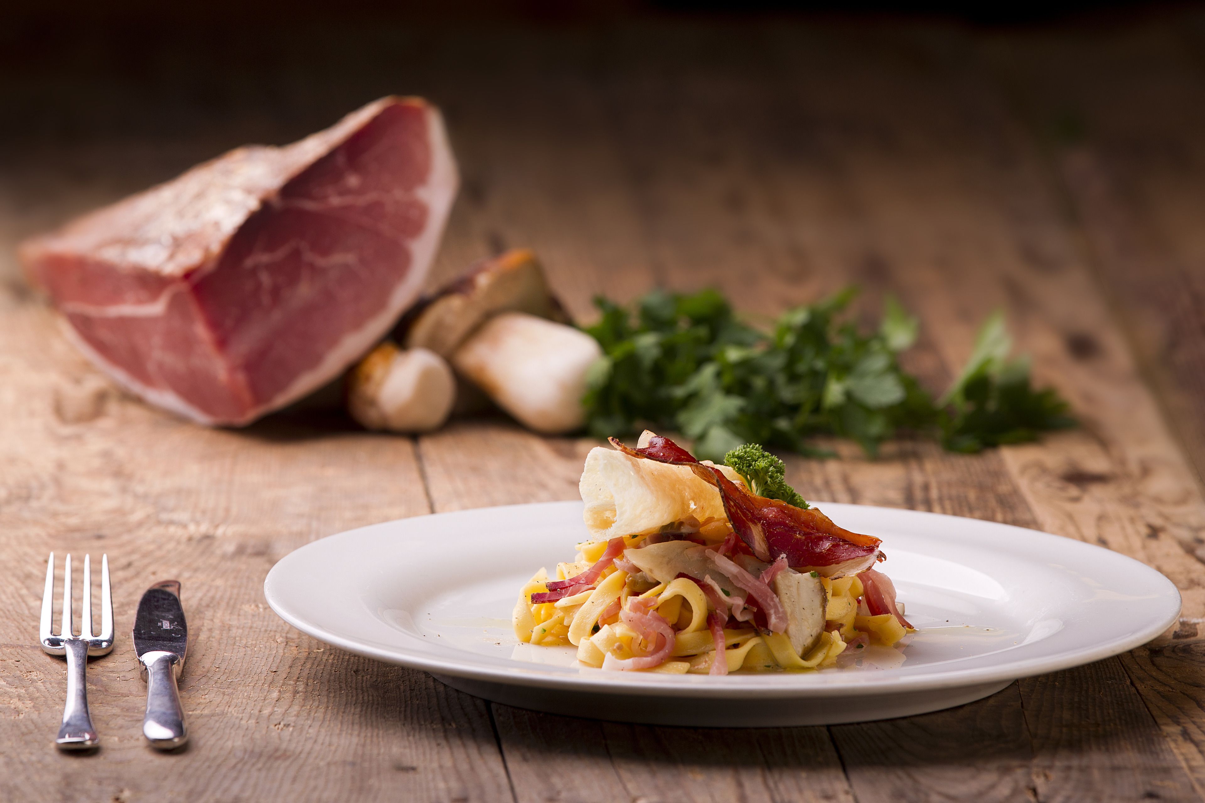 A plate of pasta with bacon and onions, a fork and knife, and a piece of ham on a wooden table.