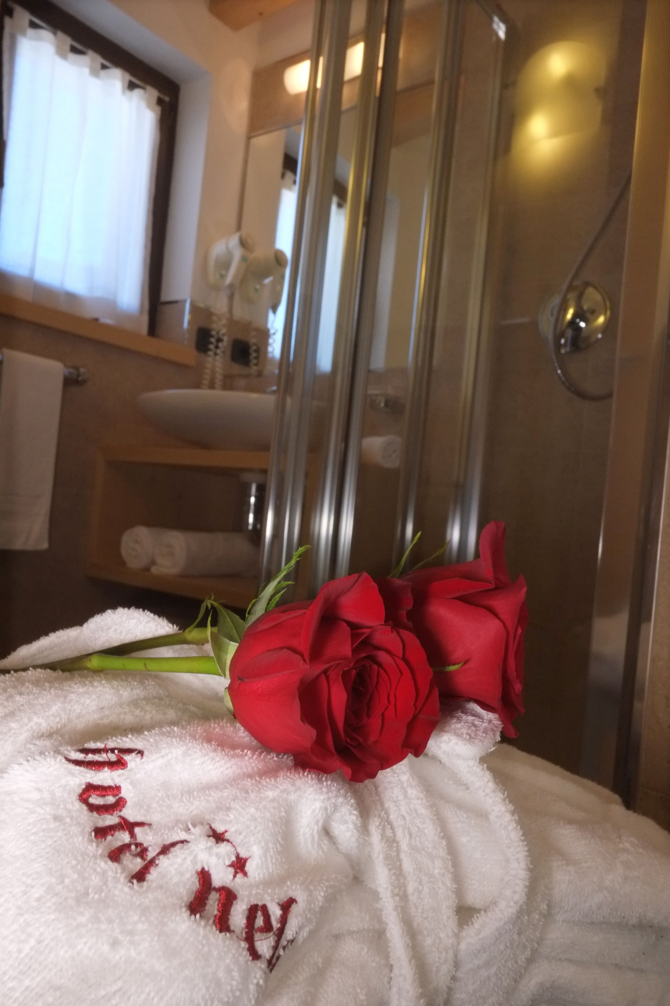Et badeværelse med en glasbrusekabine, et hvidt håndklæde med broderet tekst, og en rød rose.