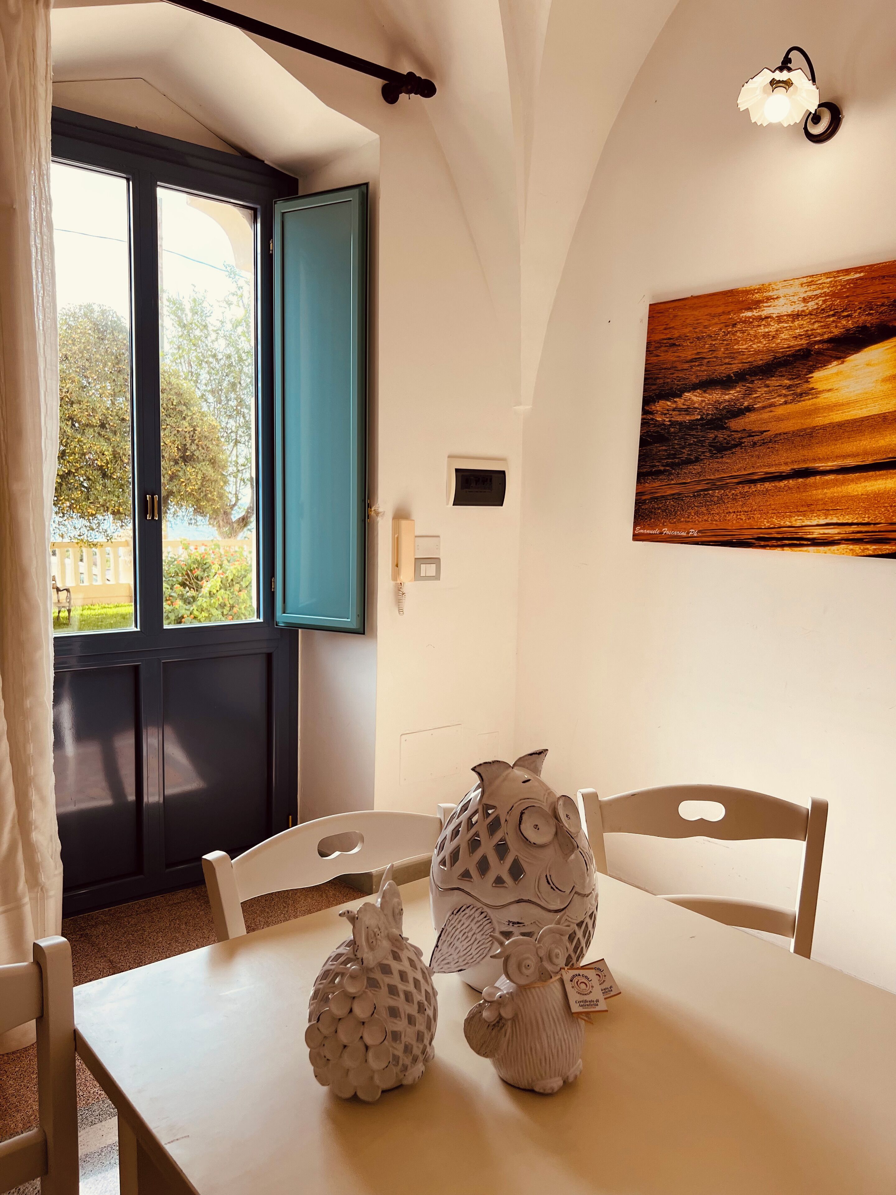 A room with a white table, two chairs, a window with blue shutters, a wall-mounted light fixture, and a painting.