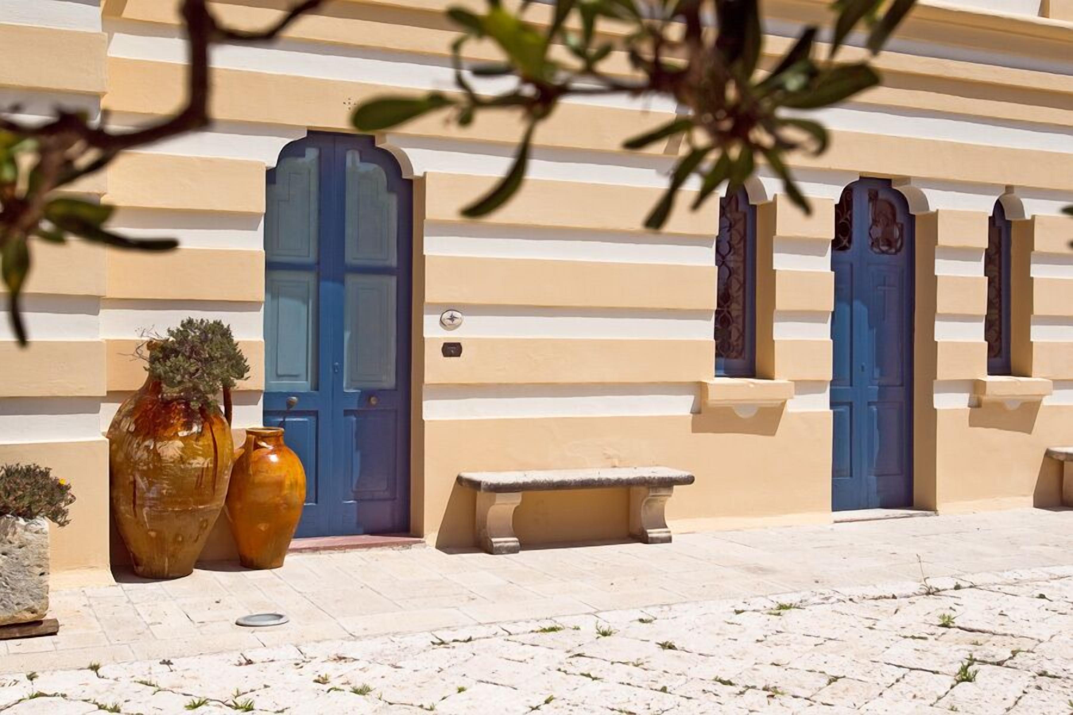 Un bâtiment à la façade beige, aux portes bleues et doté d’un banc en pierre devant l’entrée.