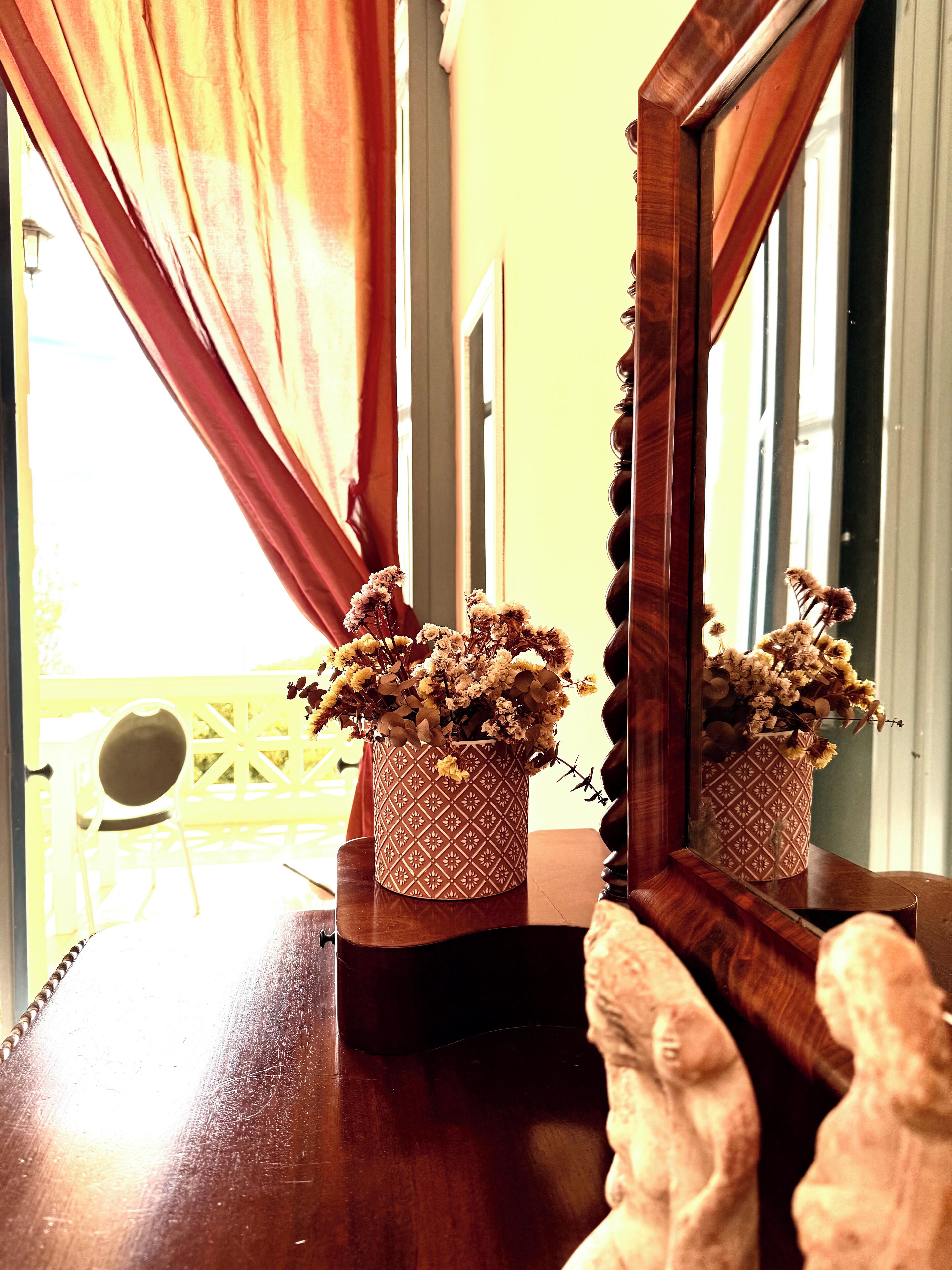 A wooden table with a patterned vase containing dried flowers, positioned in front of a mirror reflecting a window with curtains.