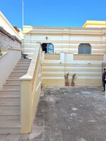 A building with a stone staircase leading to a landing, a small balcony with a potted plant, and a person standing near the entrance.