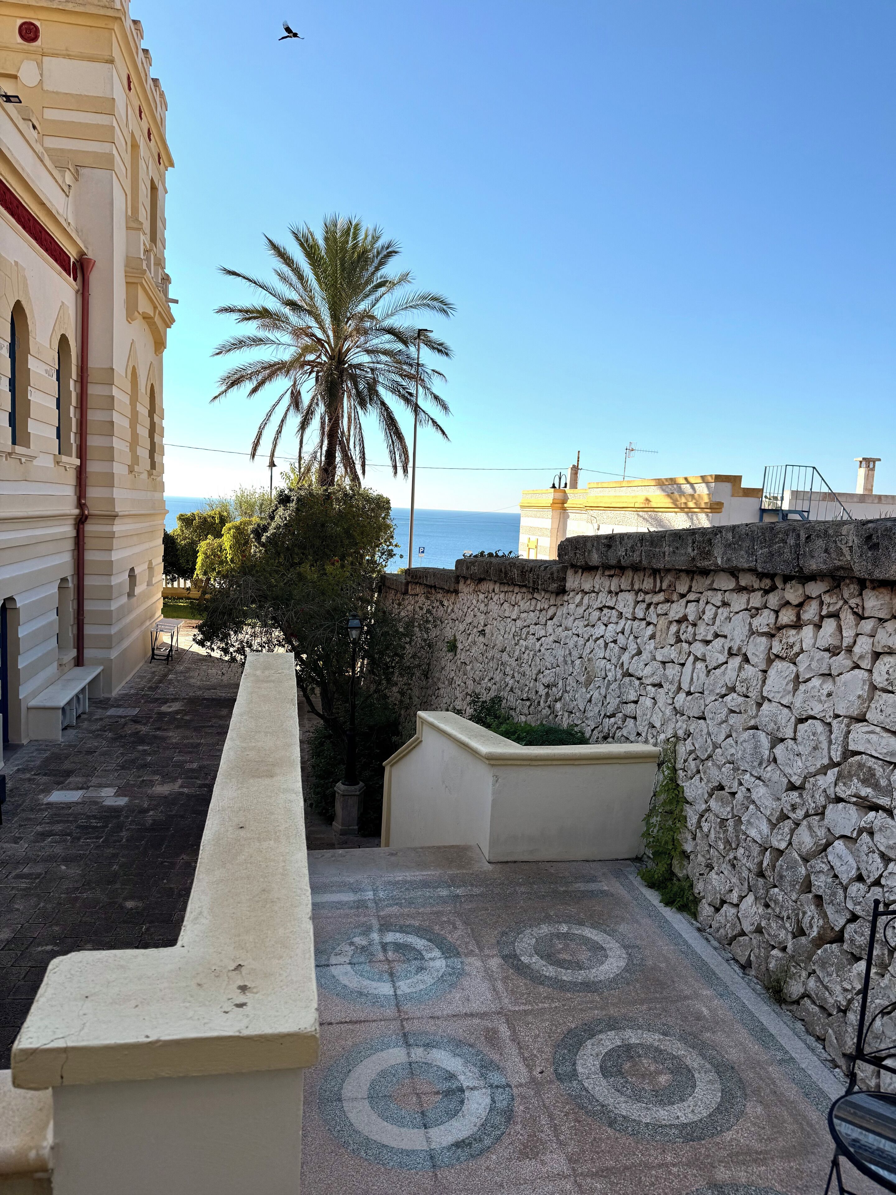 A courtyard with a stone wall, a palm tree, and a view of the sea.