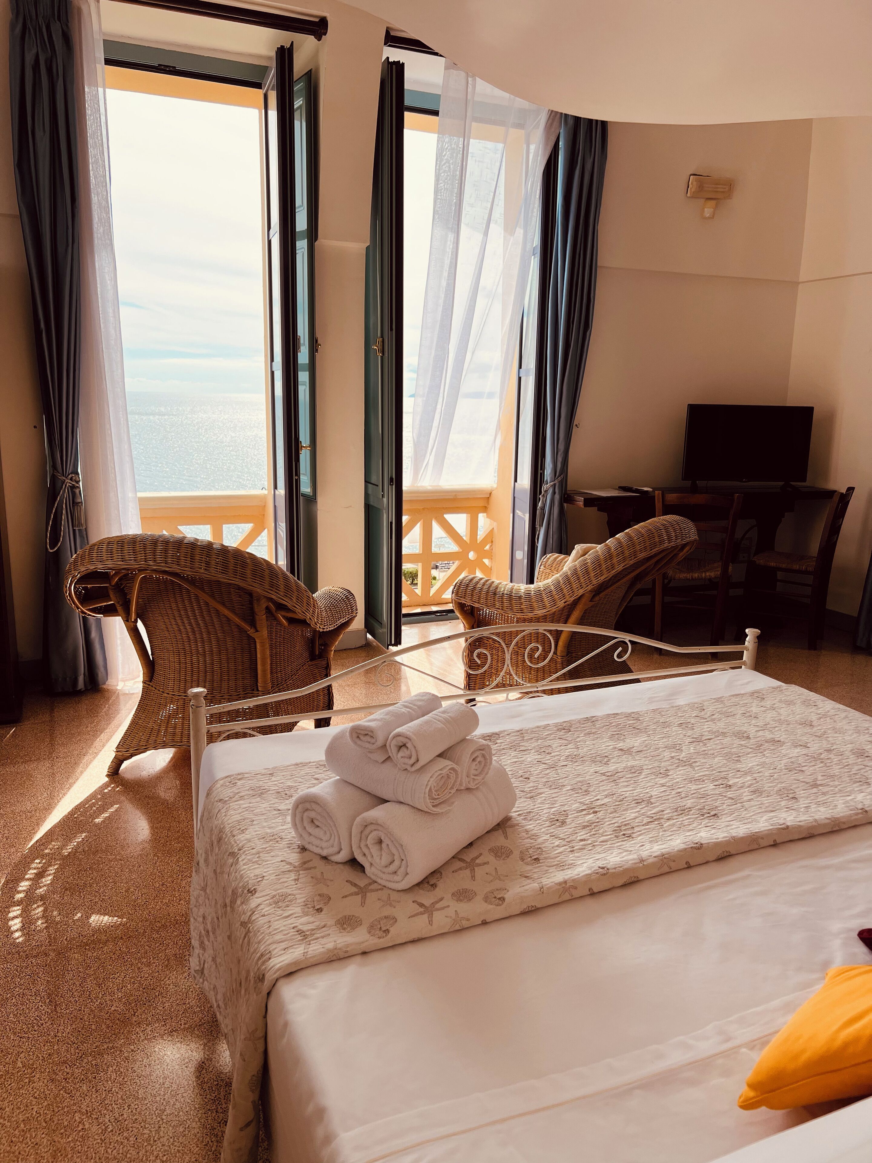 A hotel room with a bed, two wicker chairs, a television, and a view of the sea through open doors.