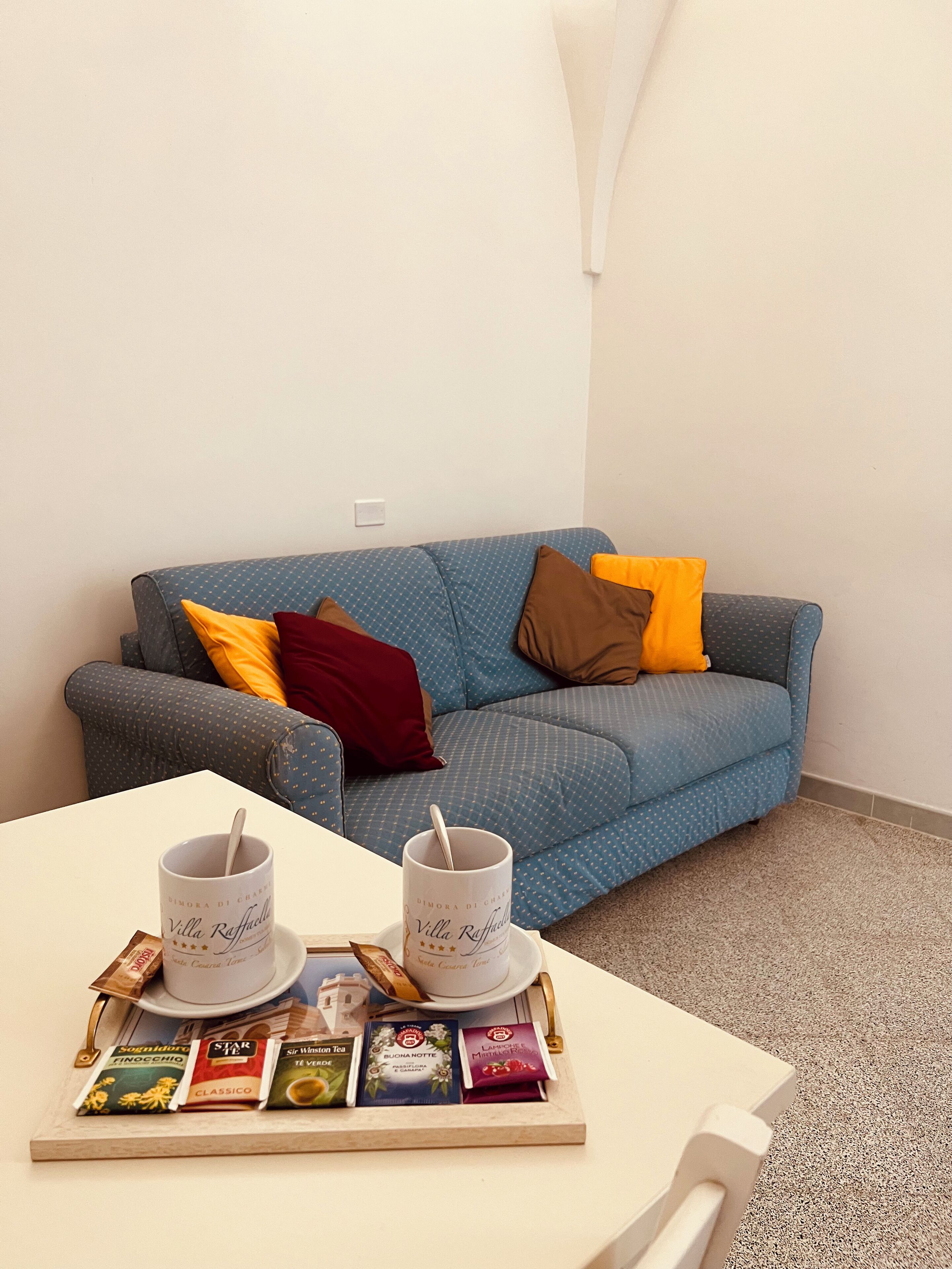 A cozy room with a blue patterned sofa, a white table with two cups and saucers, and a tray of assorted tea bags.