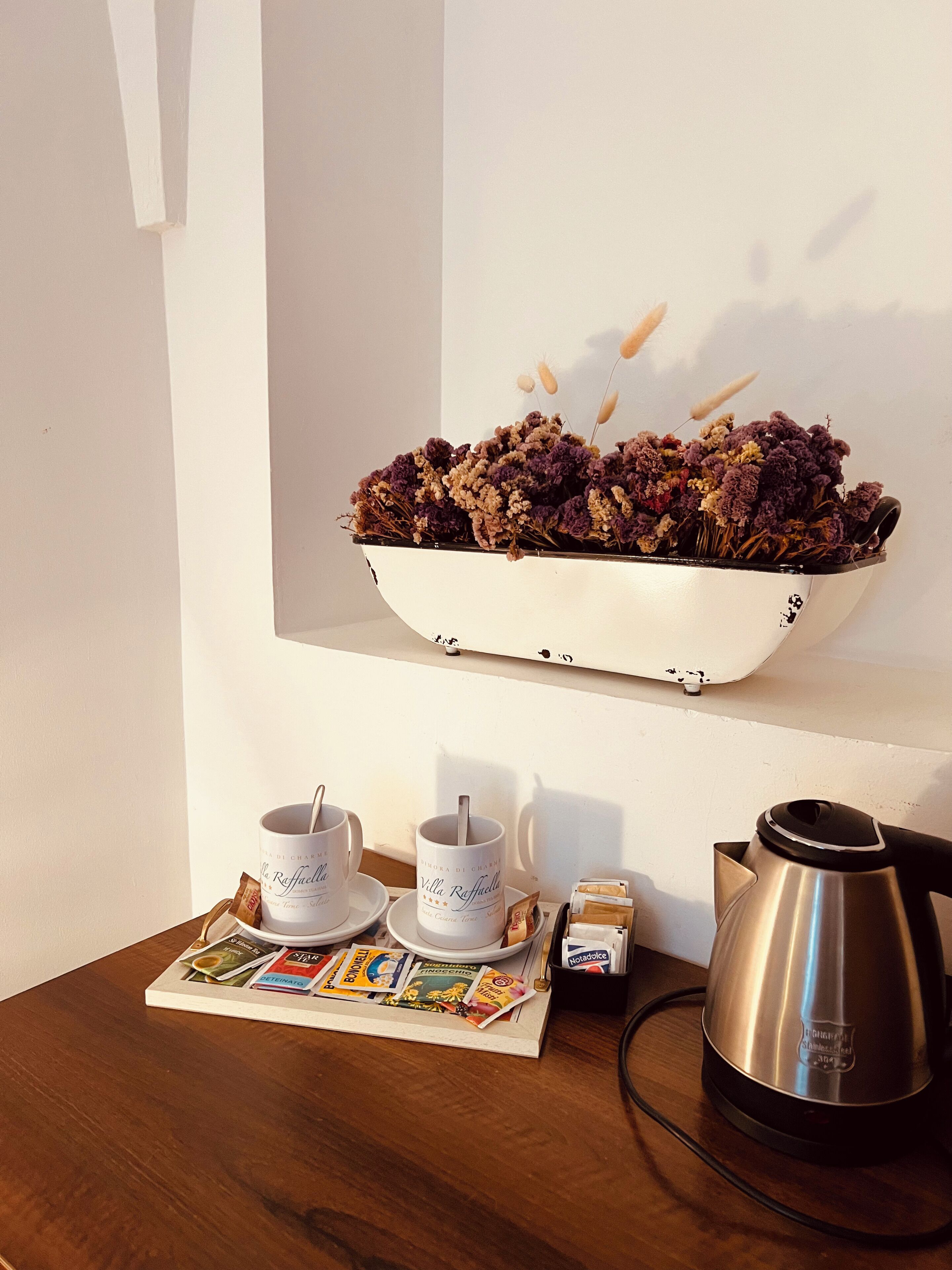 A wooden table with two white cups, a tray of dried flowers, a small box, and a stainless steel kettle.