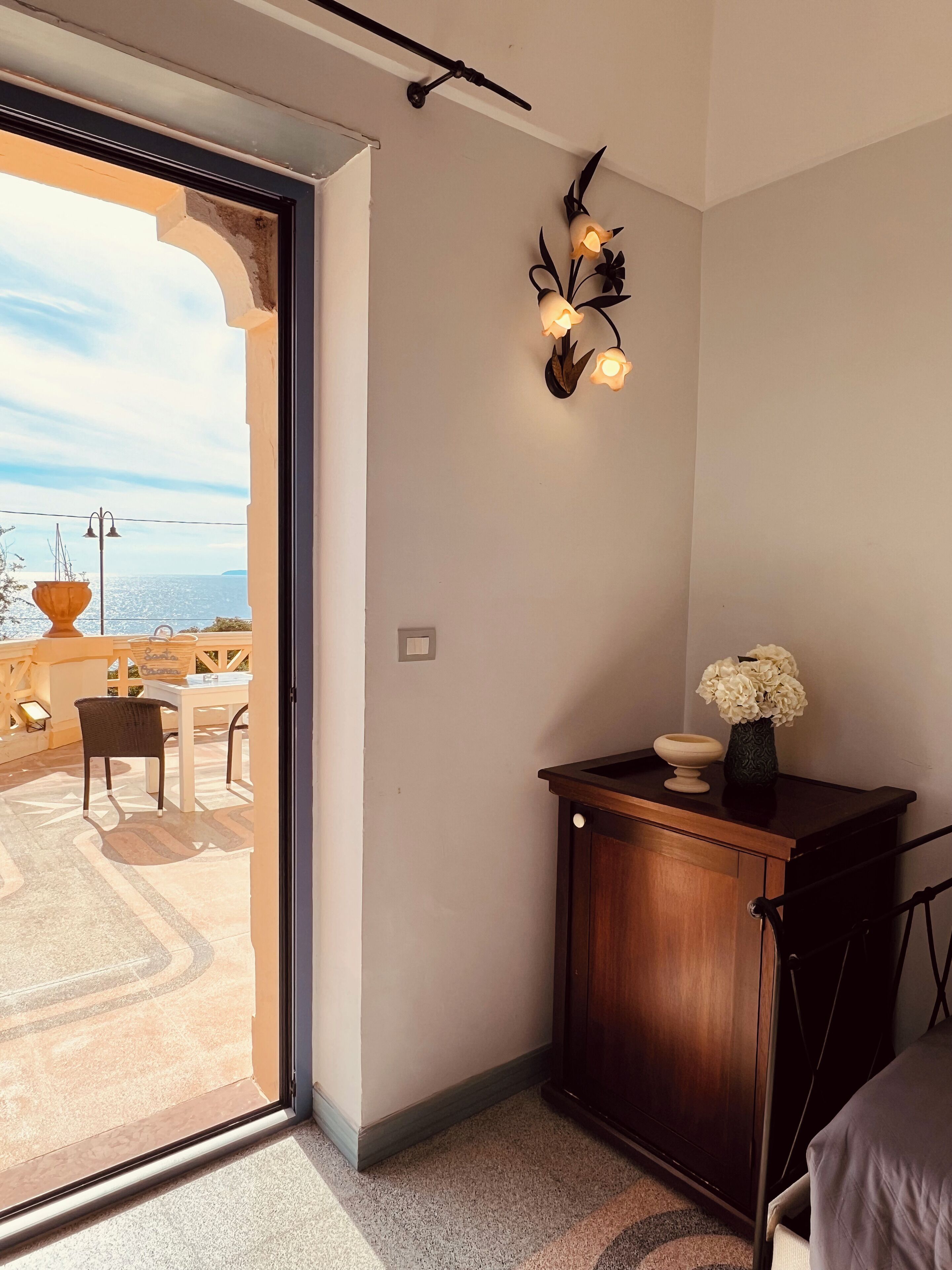 A room with a view of a terrace, a wooden cabinet with a vase, and a wall-mounted light fixture.