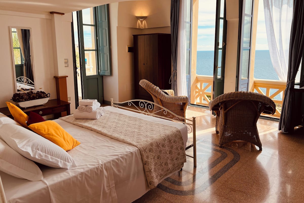 A hotel room with a bed, a chair, a small table, a mirror, and a view of the sea.