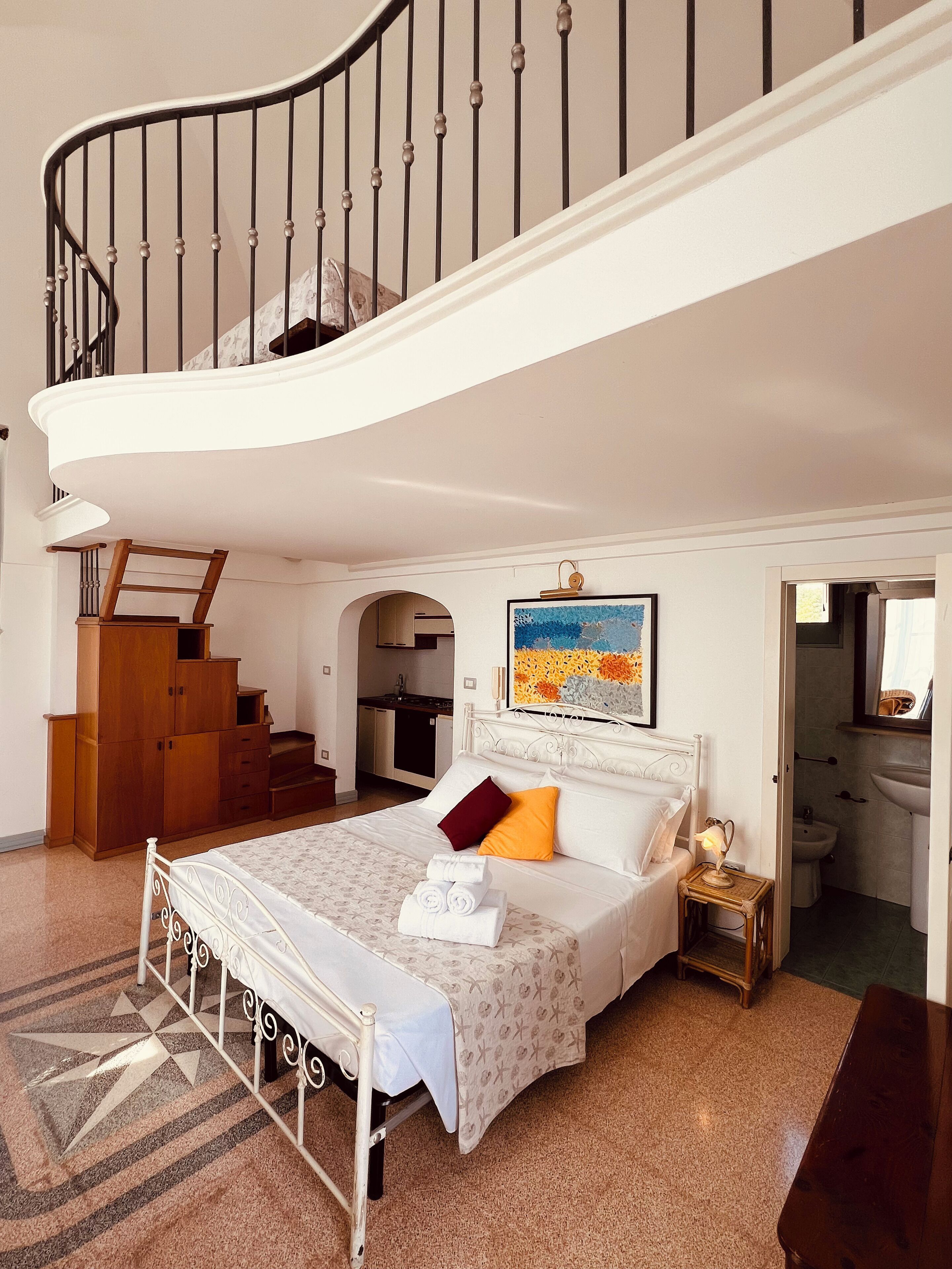 A bedroom with a curved staircase, a bed with a headboard, a bedside table with a lamp, and a view into the kitchen and bathroom.