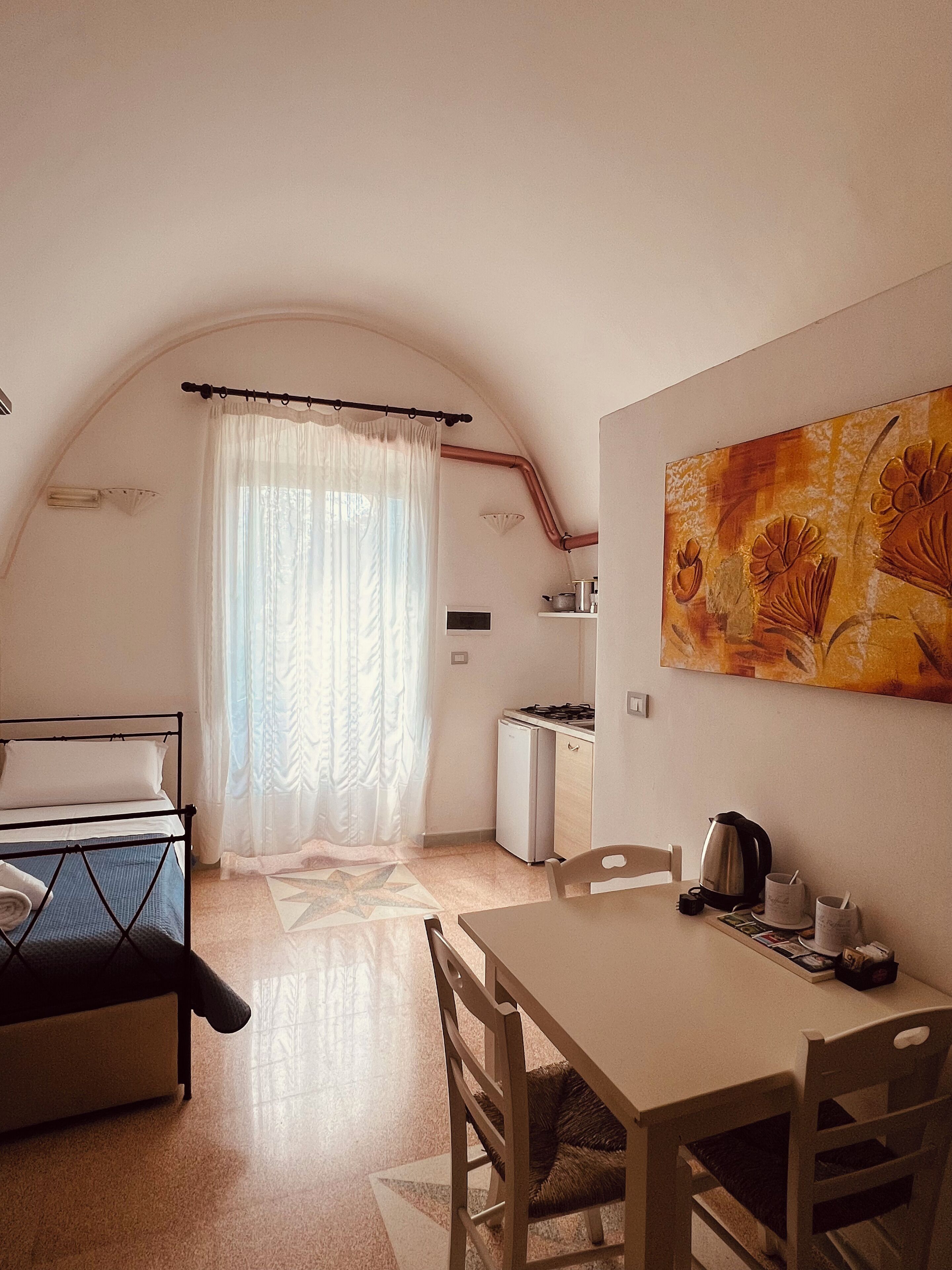 Een knus ingerichte kamer met een bed, een klein keukentje, een eettafel en een schilderij aan de muur.