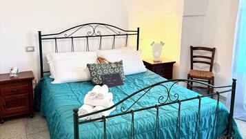 Un lit bien fait, recouvert d’une couvre-lit turquoise, avec une tête de lit en métal noir et une table de chevet surmontée d’une lampe.