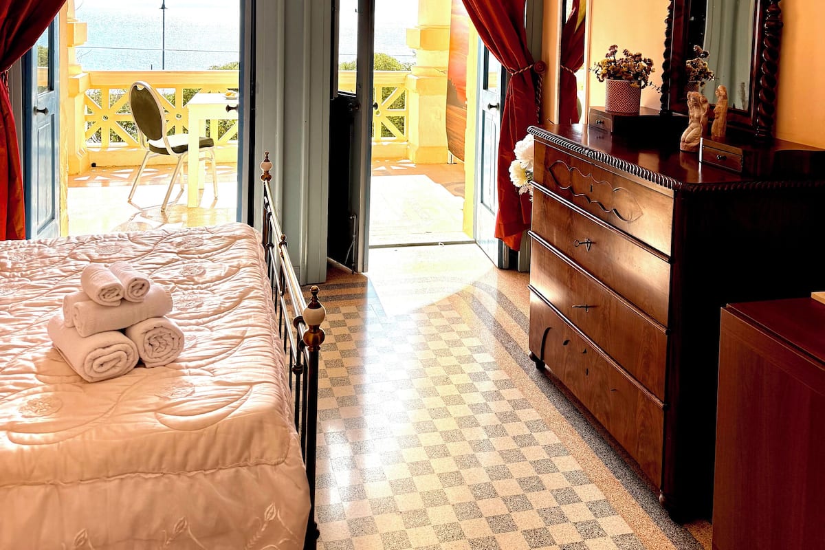 A hotel room with a bed, a dresser, a mirror, and a view of the sea through a balcony door.
