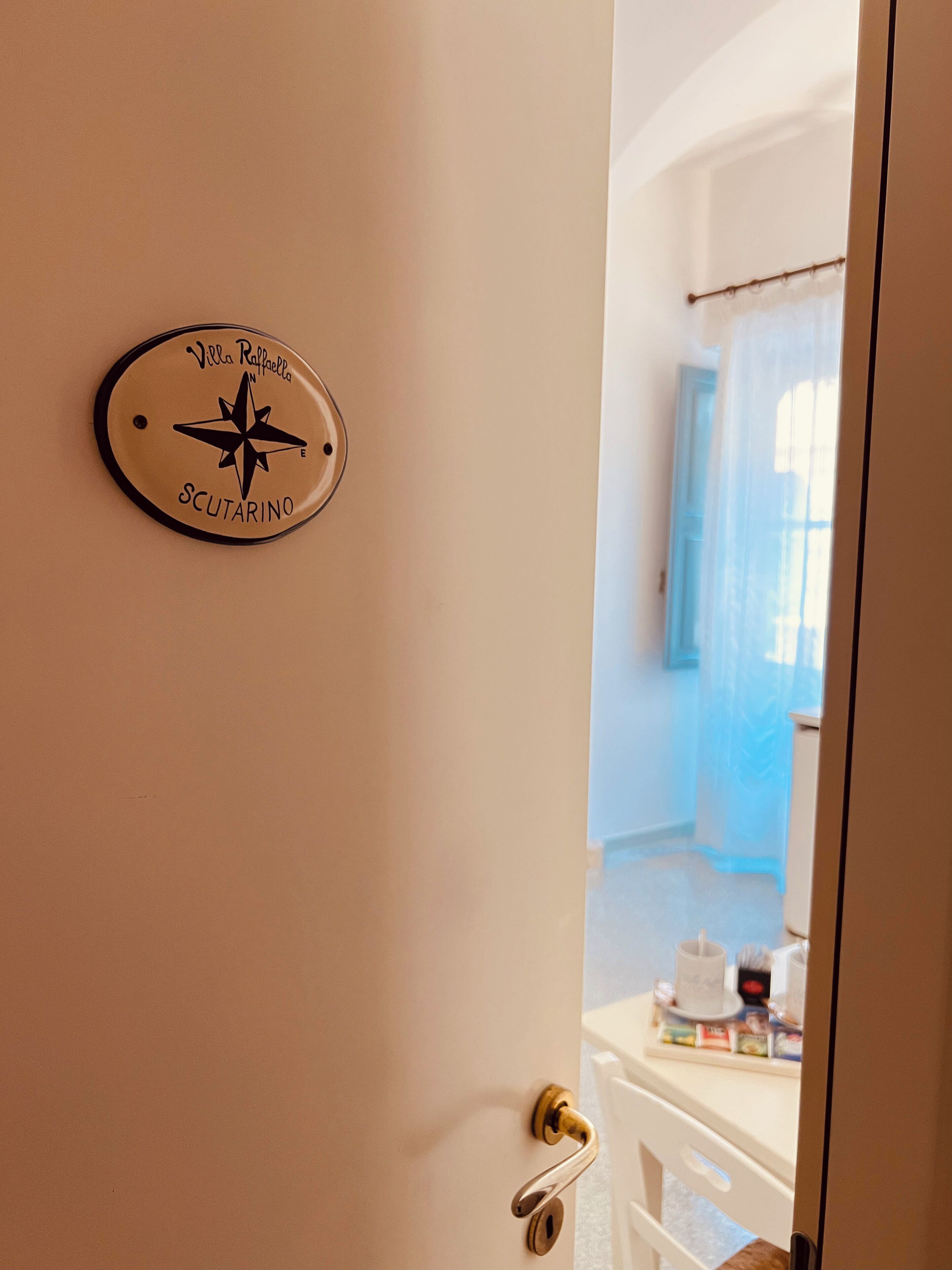 A door with a compass sign reading "Villa Raffella SCUTARINO" and a glimpse of a room with a blue curtain and a white cabinet.