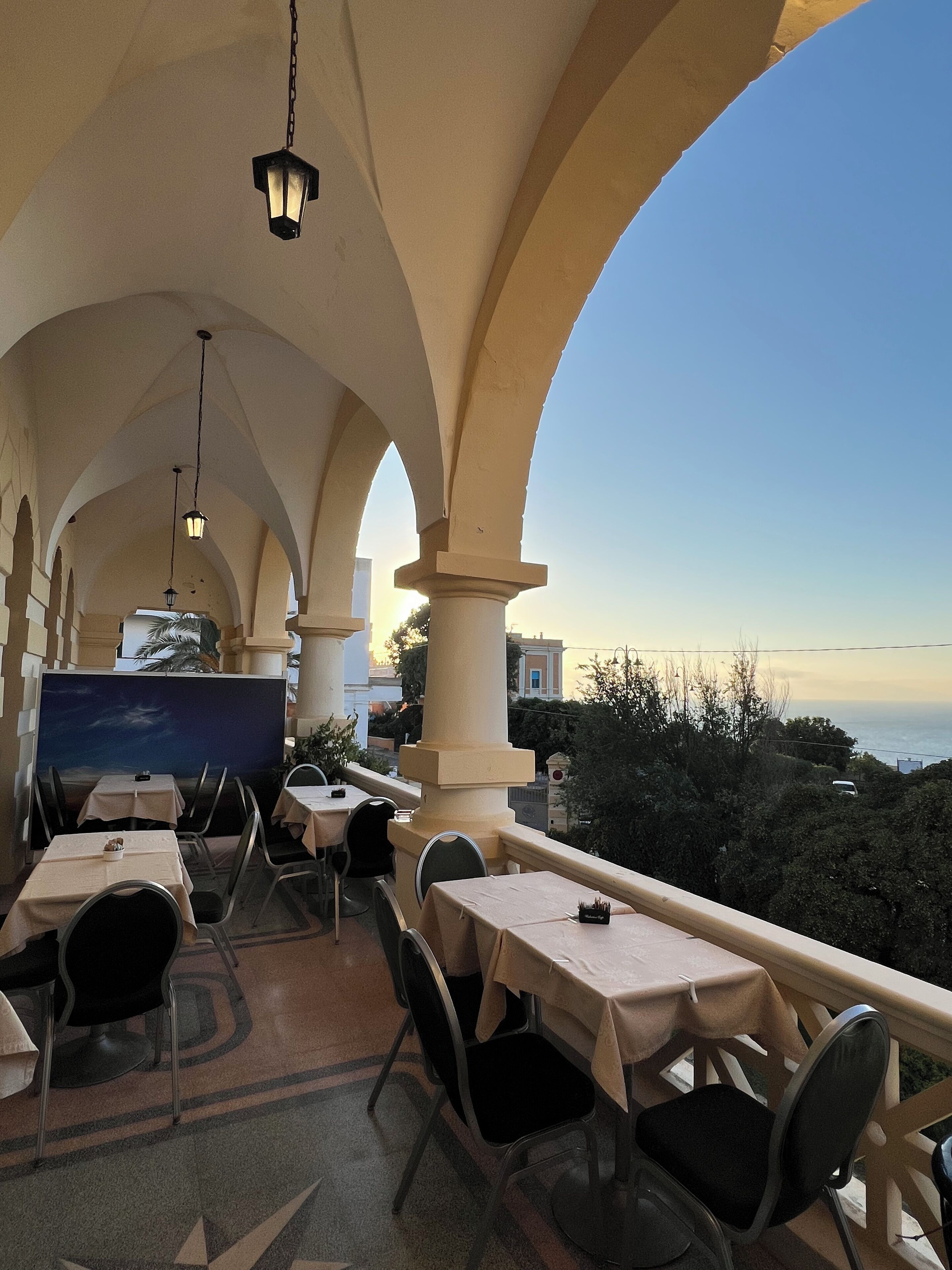An outdoor dining area with tables and chairs, a view of the sea, and a sunset in the background.