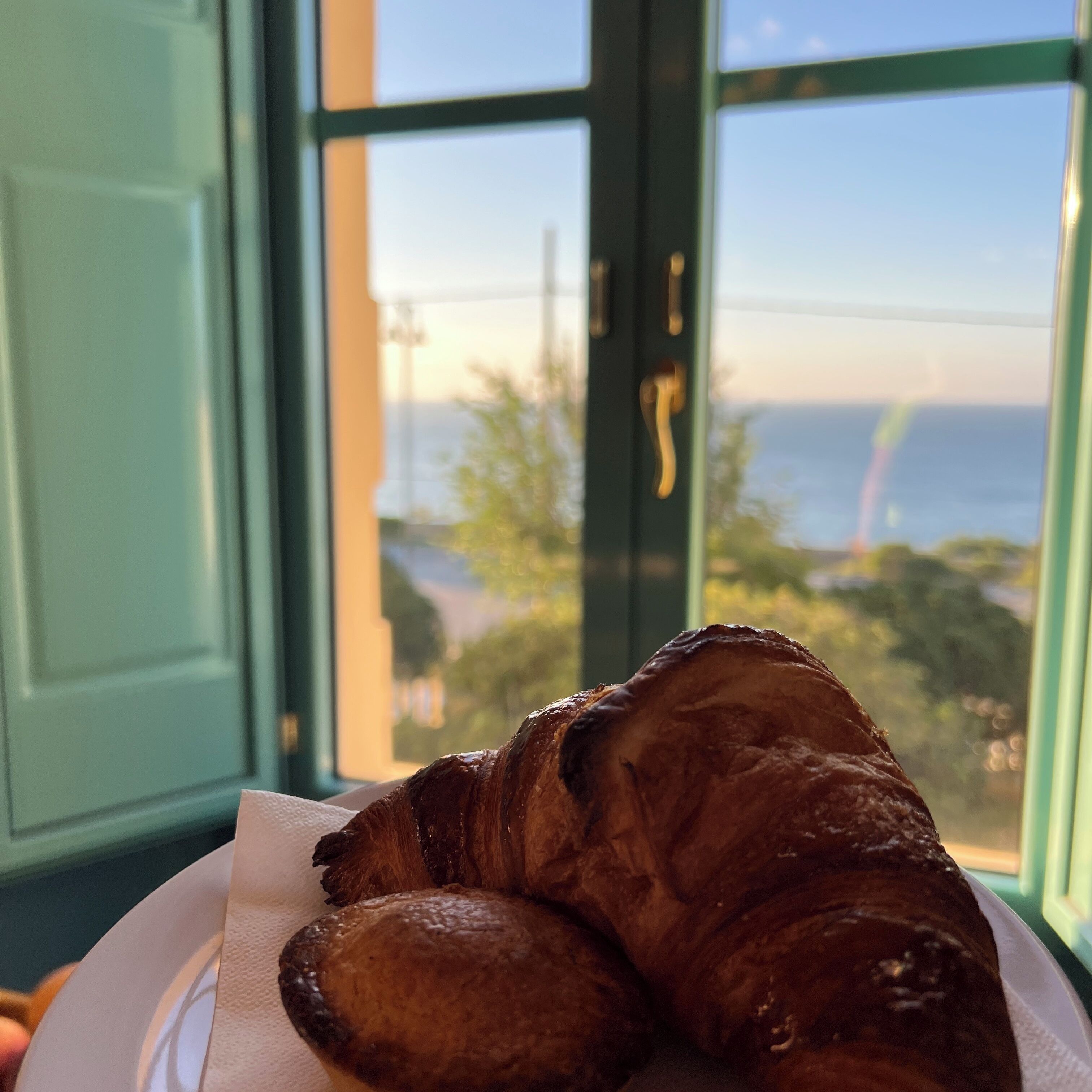 Un croissant sur une assiette blanche, accompagné d’une serviette, est posé sur une table devant une fenêtre donnant sur la mer et la verdure.
