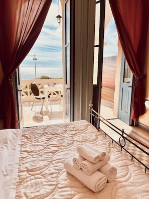A bed with a floral quilt and rolled towels, a balcony with a table and chairs, and a view of the sea.