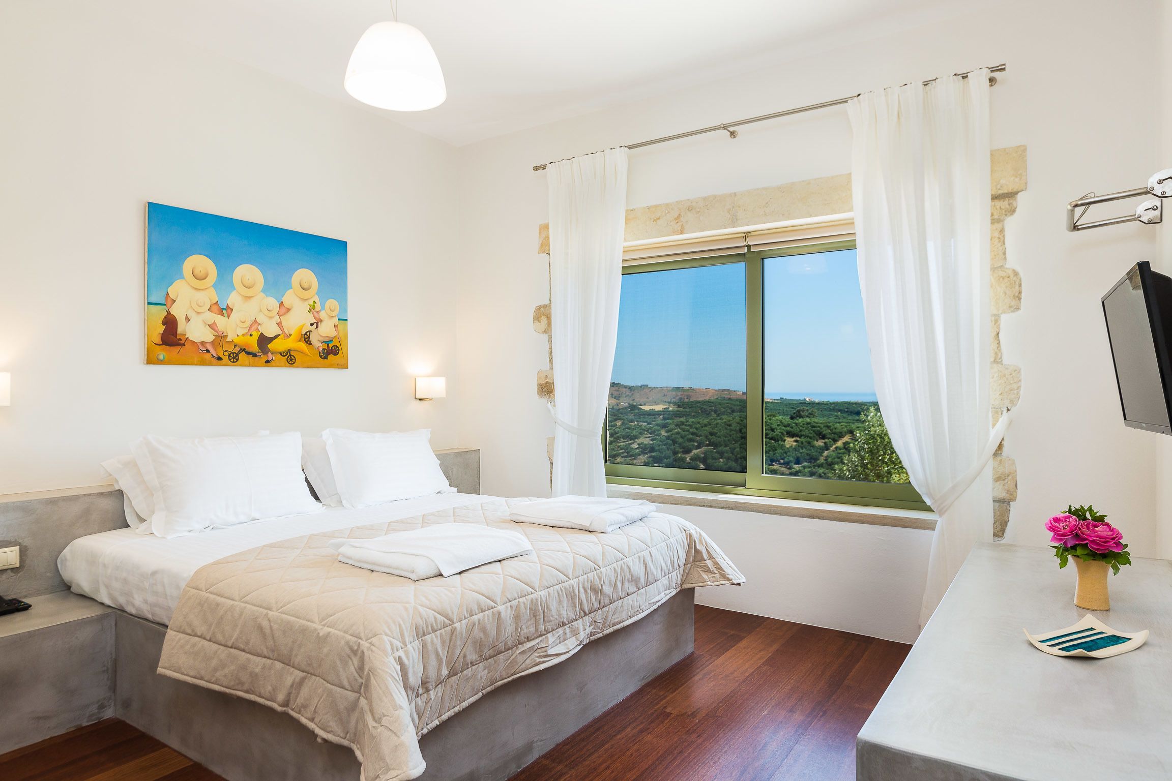 A bedroom with a large bed, a window with a view, a painting on the wall, and a bedside table with a vase of flowers.