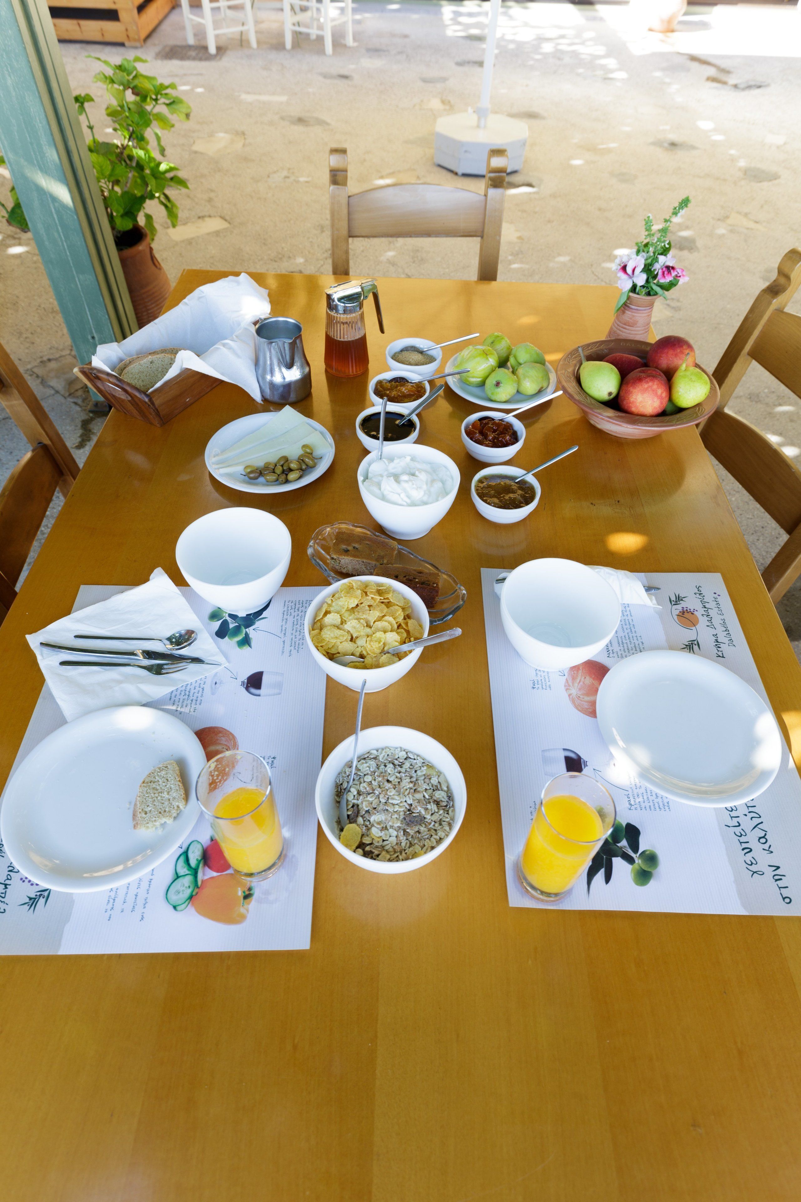 Una mesa de desayuno servida con diversos platos, entre ellos frutas, cereales y bebidas, en un entorno al aire libre y soleado.
