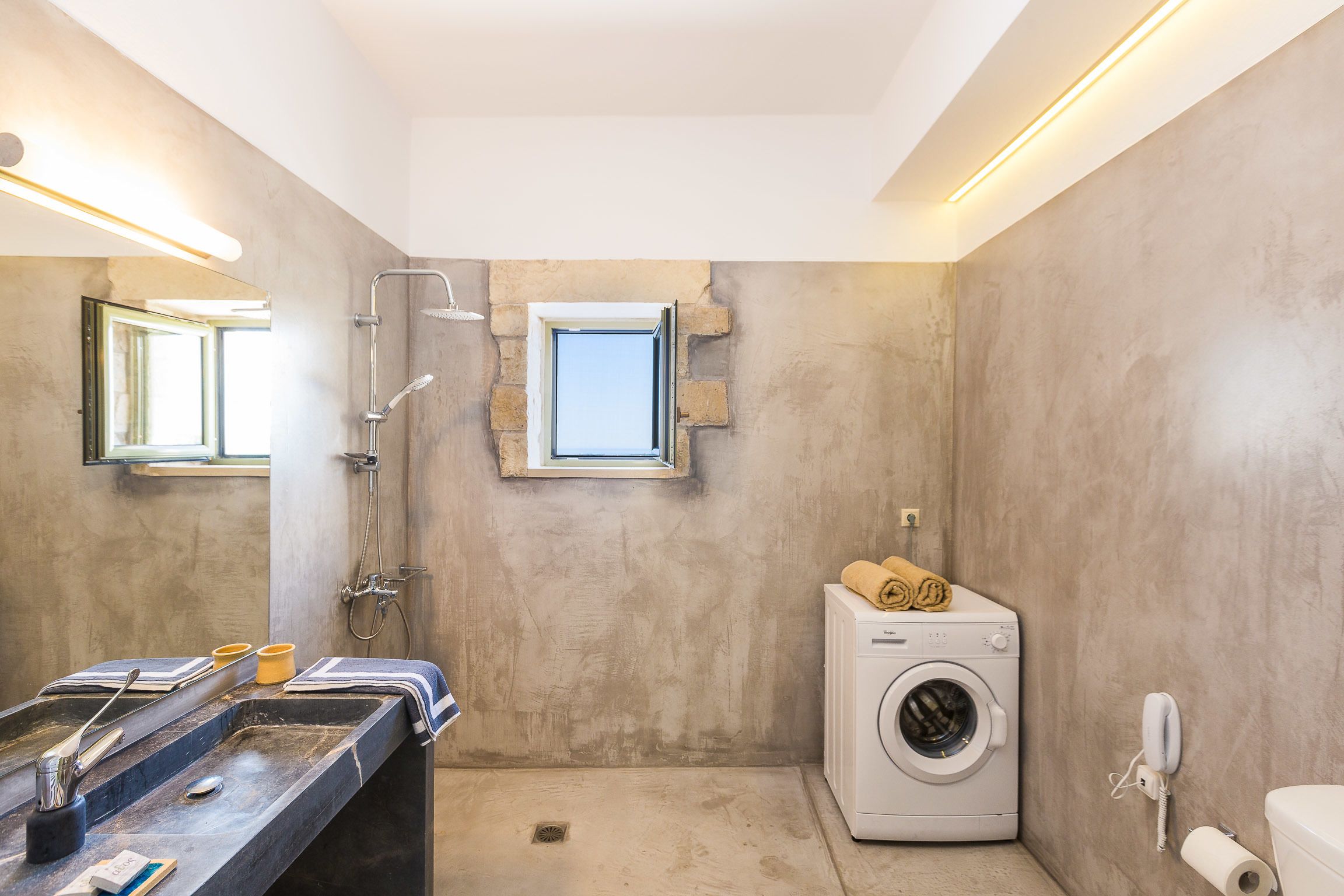 Un baño con lavamanos grande, lavadora, ducha y una ventana pequeña.