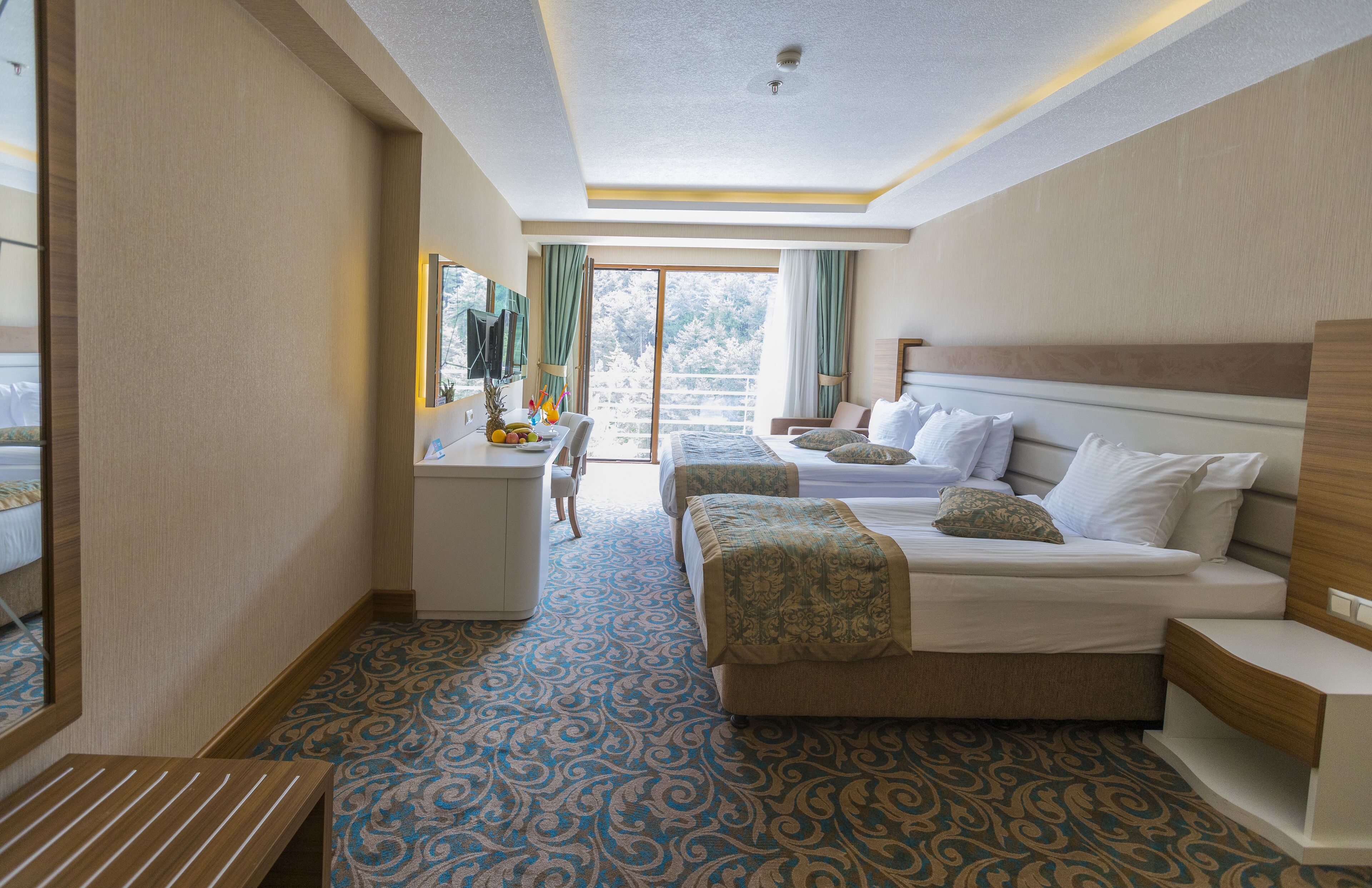 A hotel room with a large bed, a smaller bed, a desk, and a chair. There is a window with curtains, and the room has a patterned carpet.