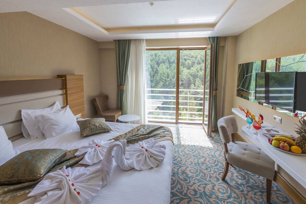 Een hotelkamer met een groot bed, een bureau met een schaal fruit, een stoel en een balkon met uitzicht op bomen.