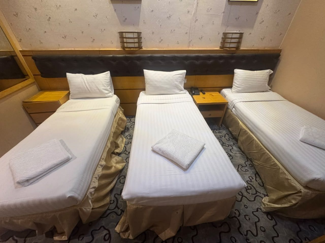 Habitación de hotel con tres camas individuales, cada una con una colcha blanca y una toalla doblada con cuidado.