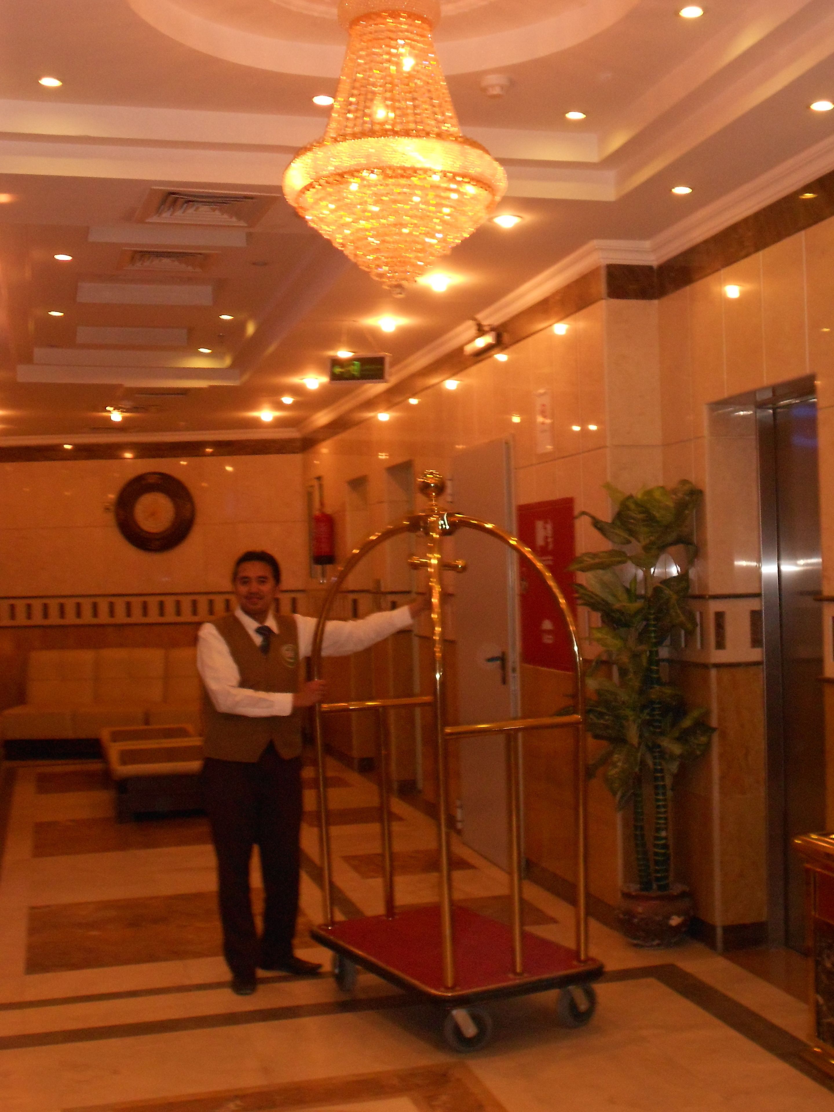 A man standing next to a gold-framed elevator with a red floor, in a lobby with a large chandelier, a clock, and a potted plant.