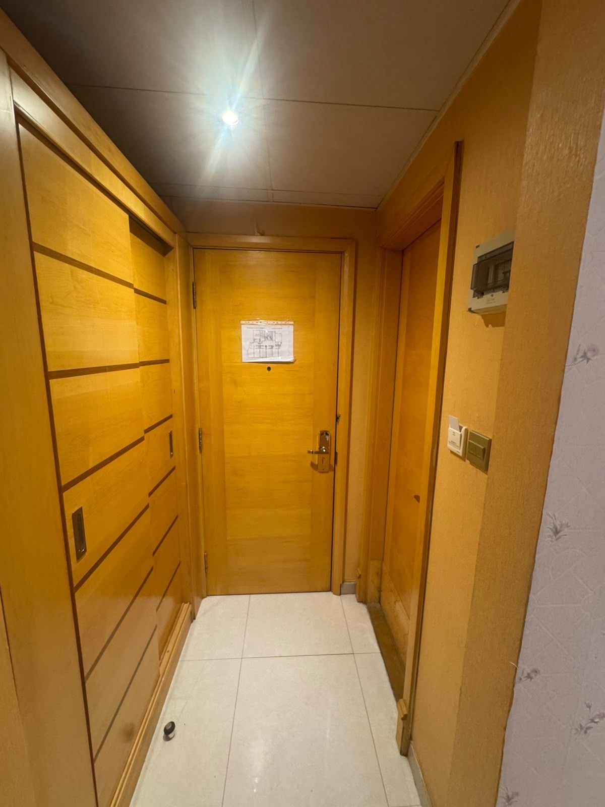 Un couloir étroit avec des portes en bois, un sol carrelé et un luminaire au plafond.