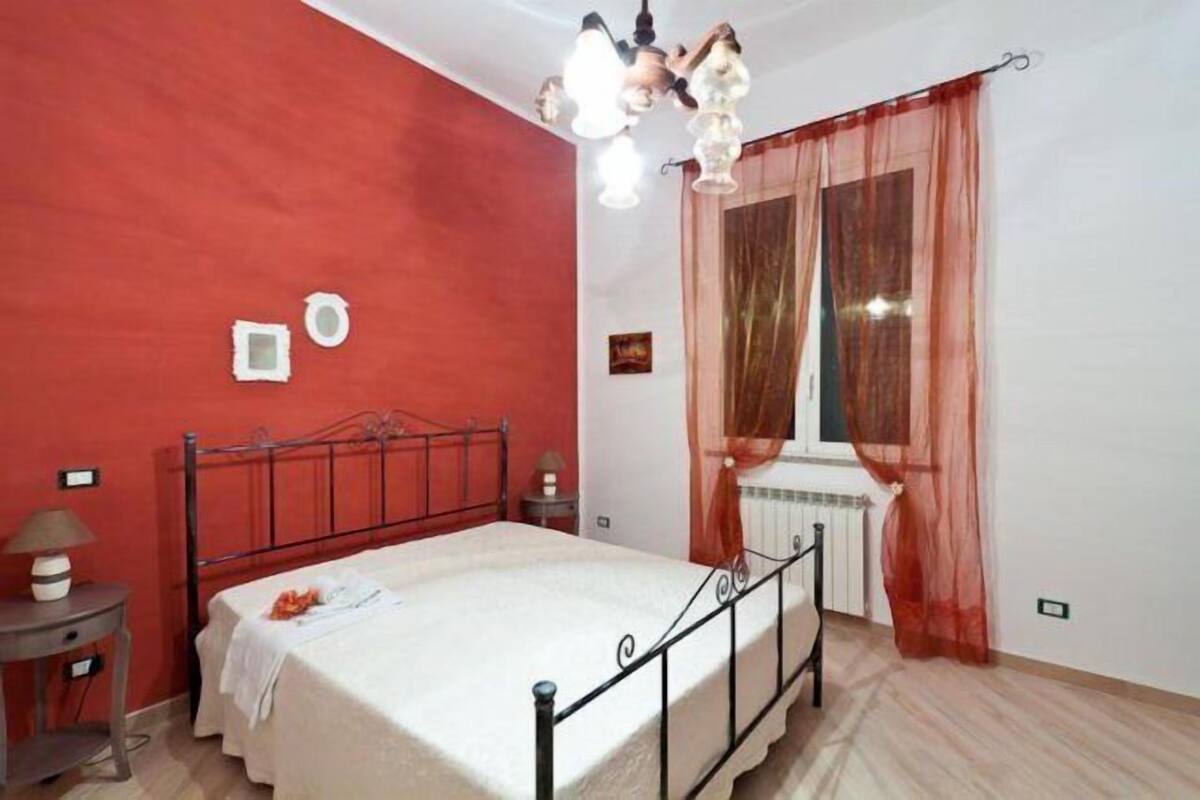 Une chambre à coucher comprenant un lit, une table de chevet, une lampe, une fenêtre avec des rideaux légers et un radiateur.