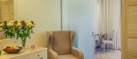 A hotel room with a beige armchair, a desk with a chair, a mirror, a vase with flowers, and a bowl of fruit.