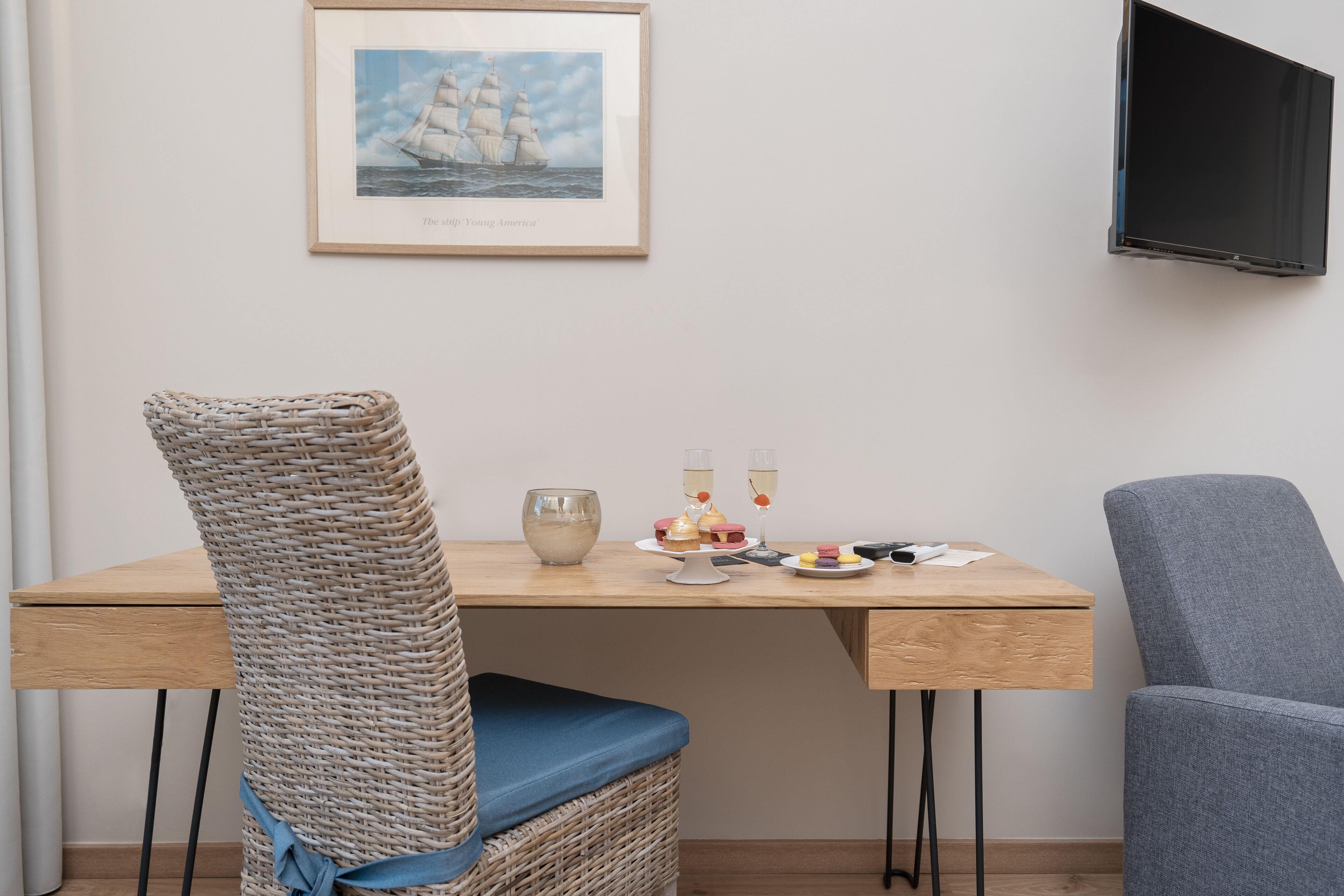 Un coin repas comprenant une chaise en osier, un fauteuil recouvert d’un tissu gris, une table en bois et un tableau encadré représentant un navire, accroché au mur.