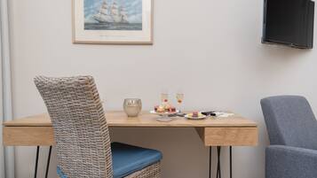 Un coin repas comprenant une chaise en osier, un fauteuil recouvert d’un tissu gris, une table en bois et un tableau encadré représentant un navire, accroché au mur.