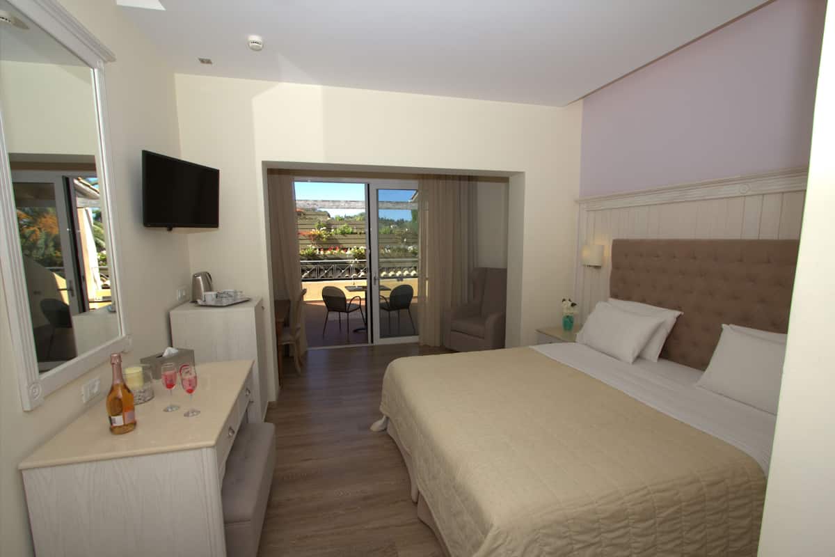 A hotel room with a bed, a TV, a desk with a bottle and glasses, a chair, and a balcony with a view.