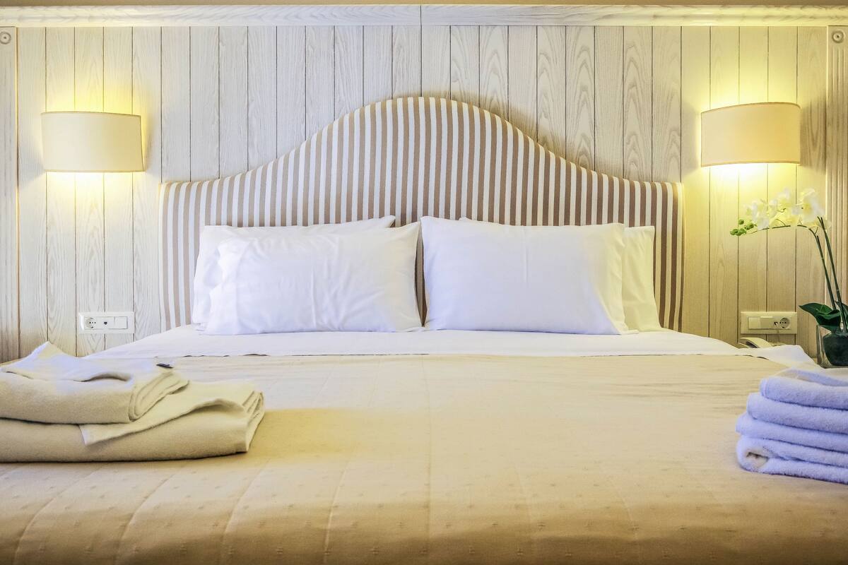 A neatly made bed with white linens and striped headboard, two bedside lamps, and a vase with flowers on the nightstand.