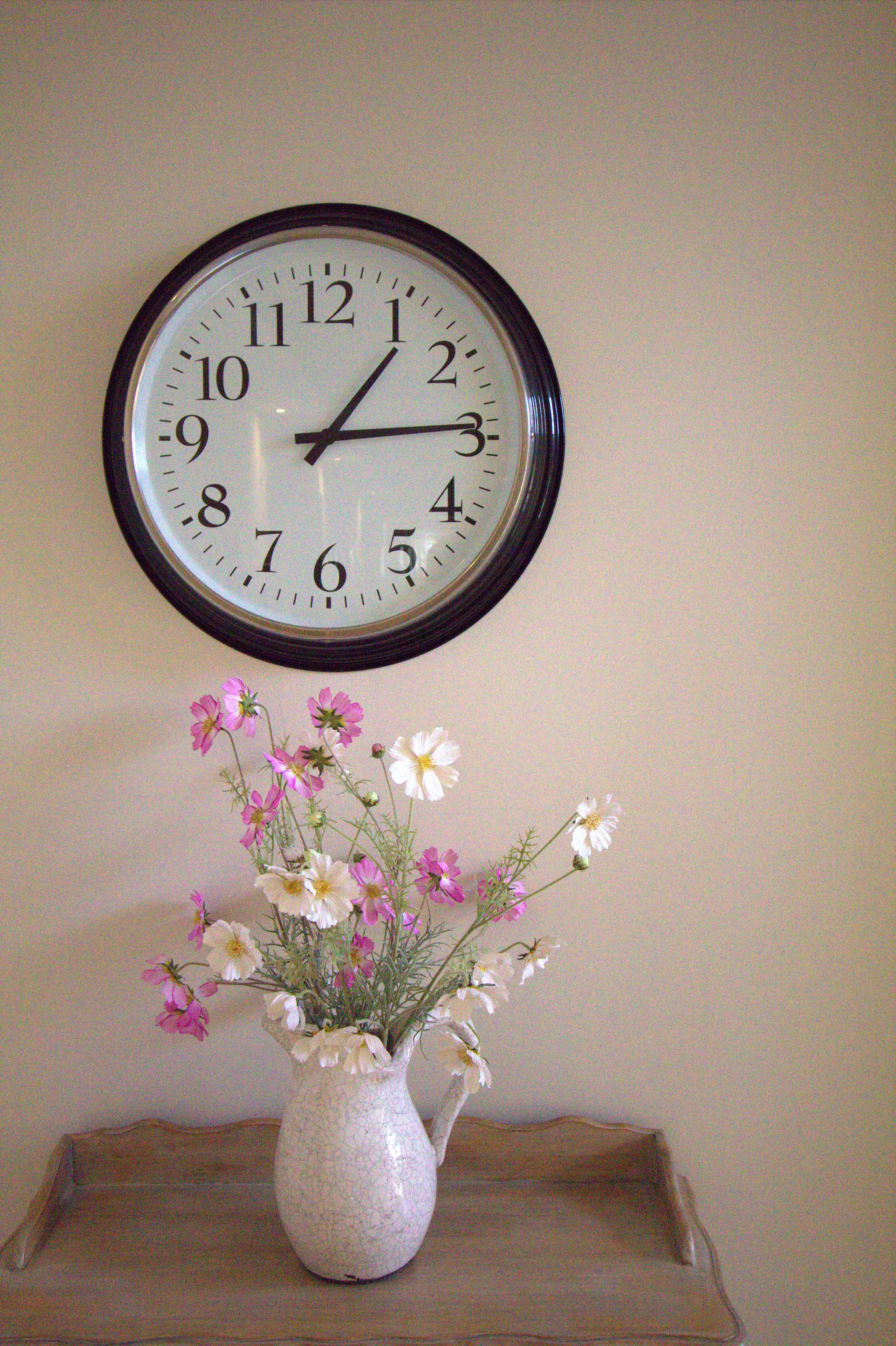 A wall clock with a white face and black numbers, and a vase with flowers on a wooden tray below it.