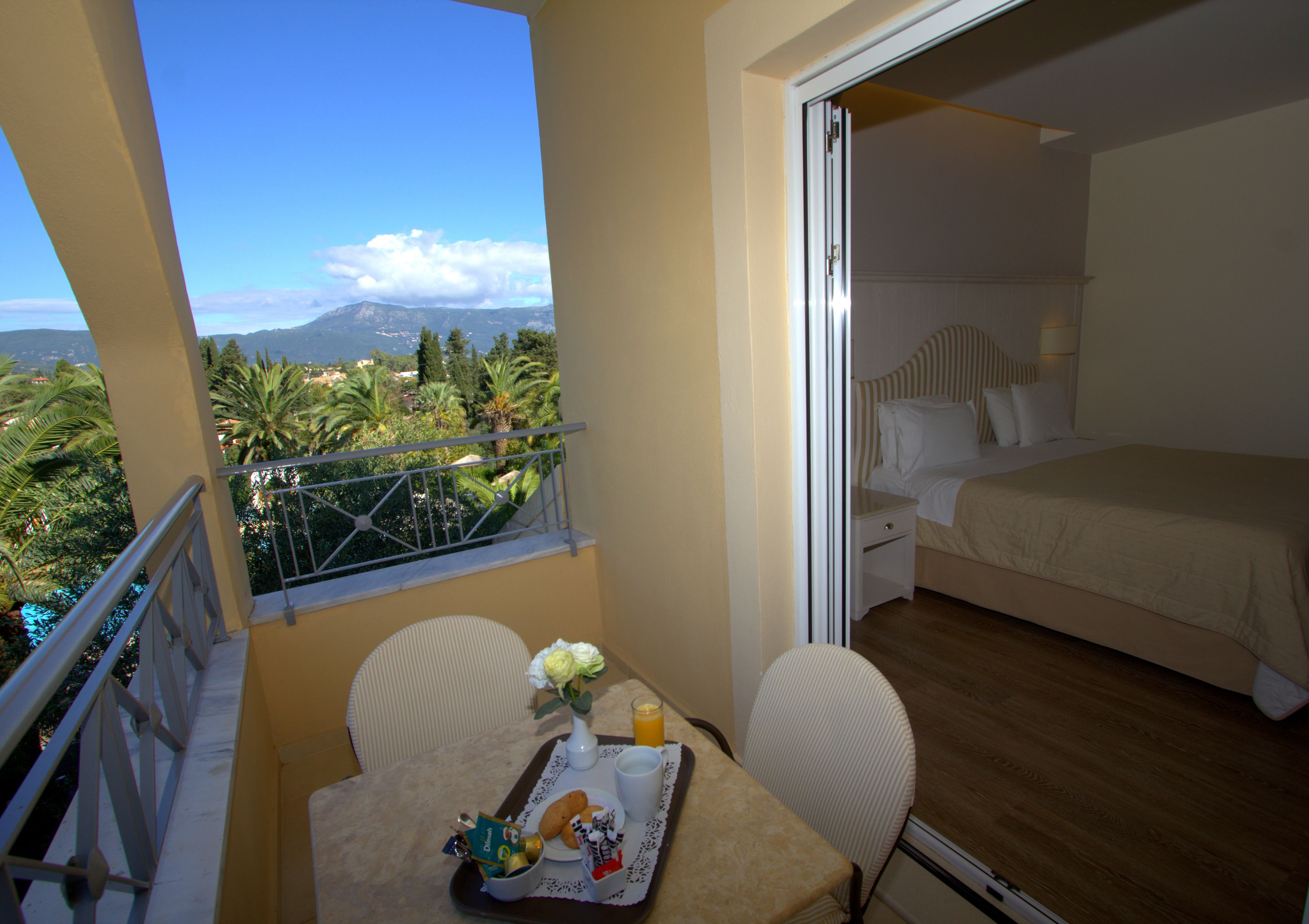 Un balcon avec une table à manger, des chaises et un plateau de petit-déjeuner. On peut apercevoir, à travers une porte ouverte, une chambre à coucher avec un lit et une table de chevet.