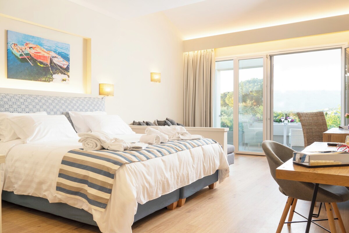 A hotel room with a bed, a desk, a chair, a window with a view, and a painting on the wall.