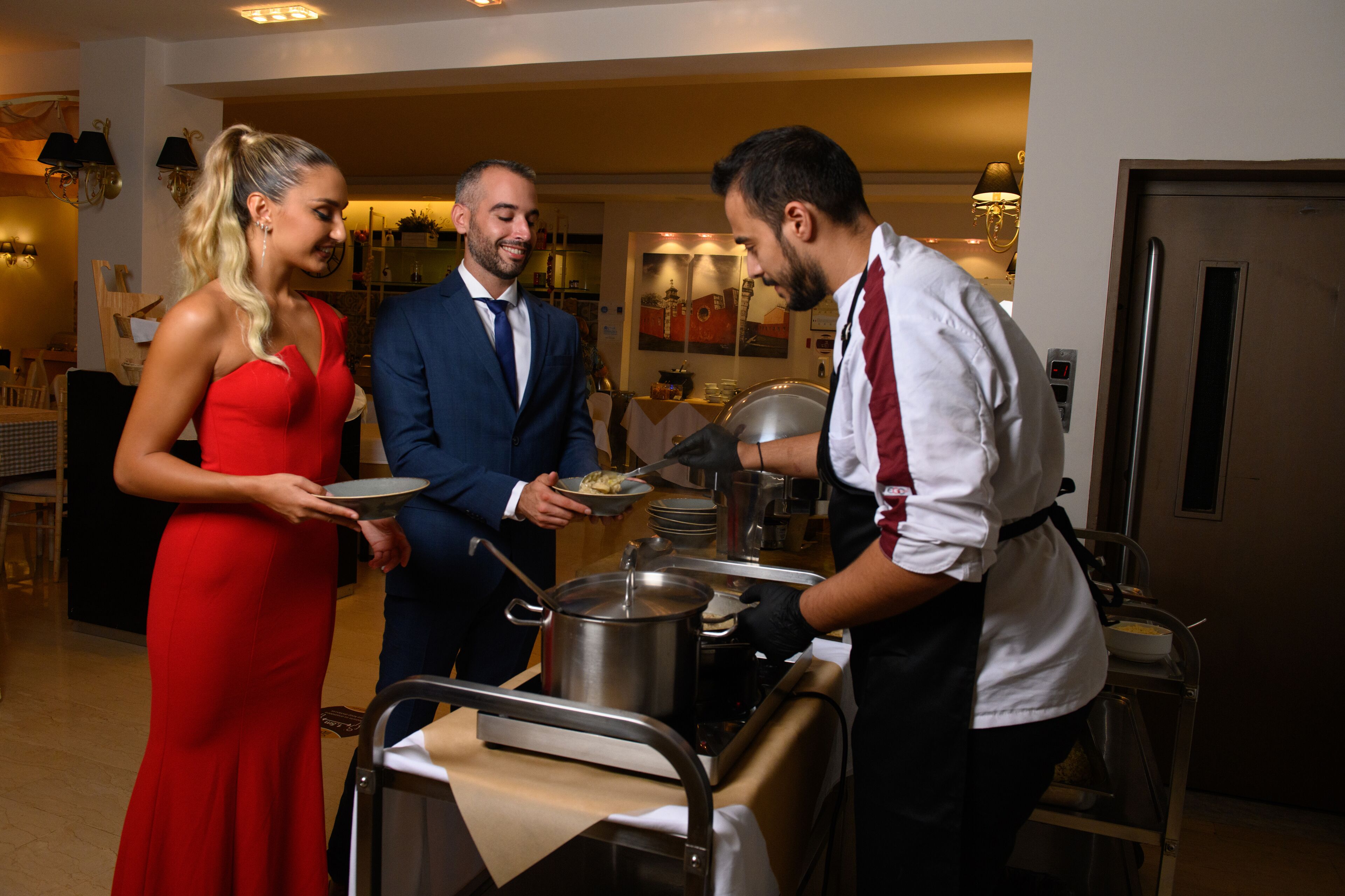 A hotel dining area with a buffet setup, including a server, a woman in a red dress, and a man in a suit.