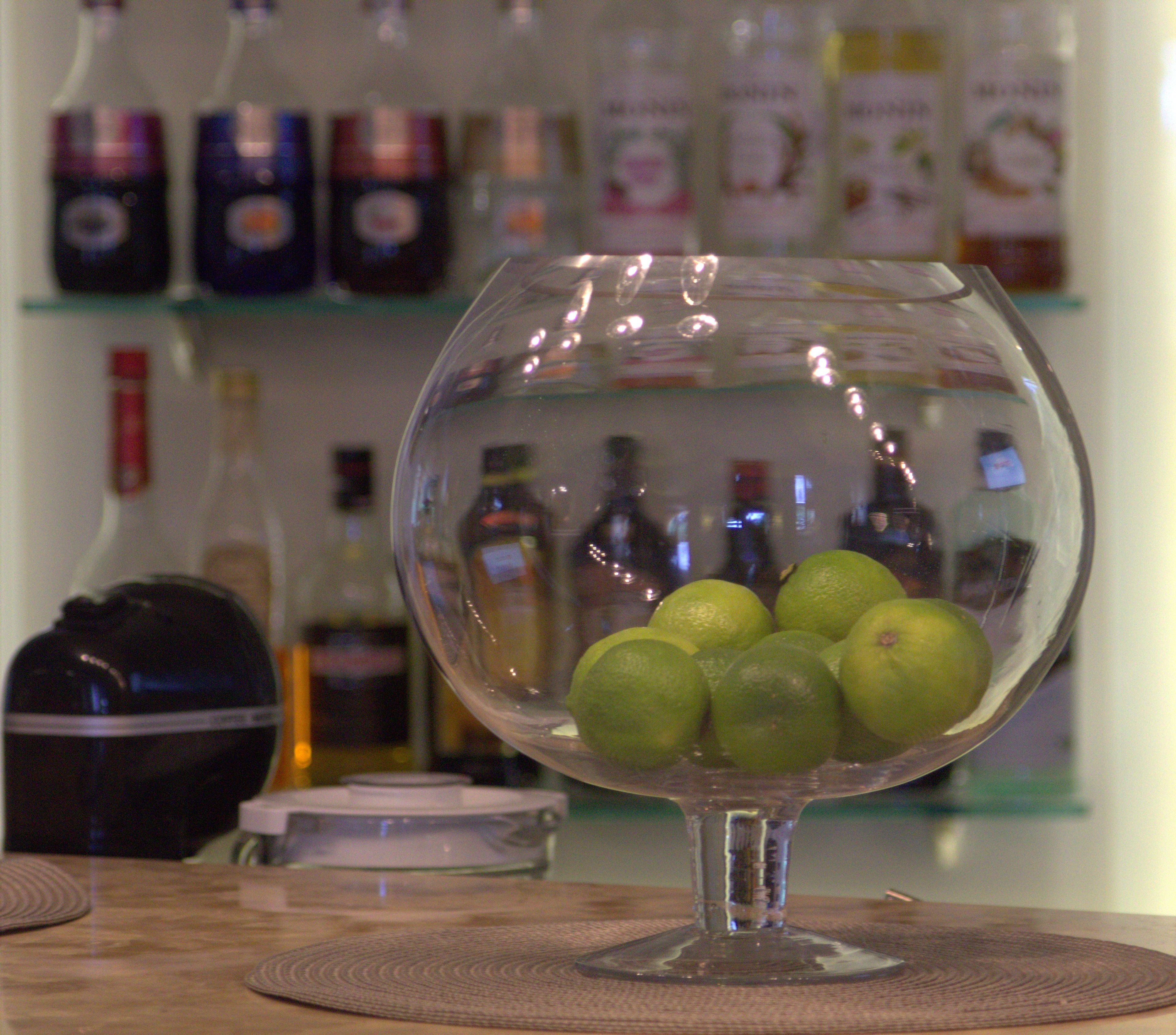 Un saladier en verre contenant des citrons verts est posé sur une table recouverte d’un set de table. En arrière-plan, on aperçoit des étagères remplies de différentes bouteilles.