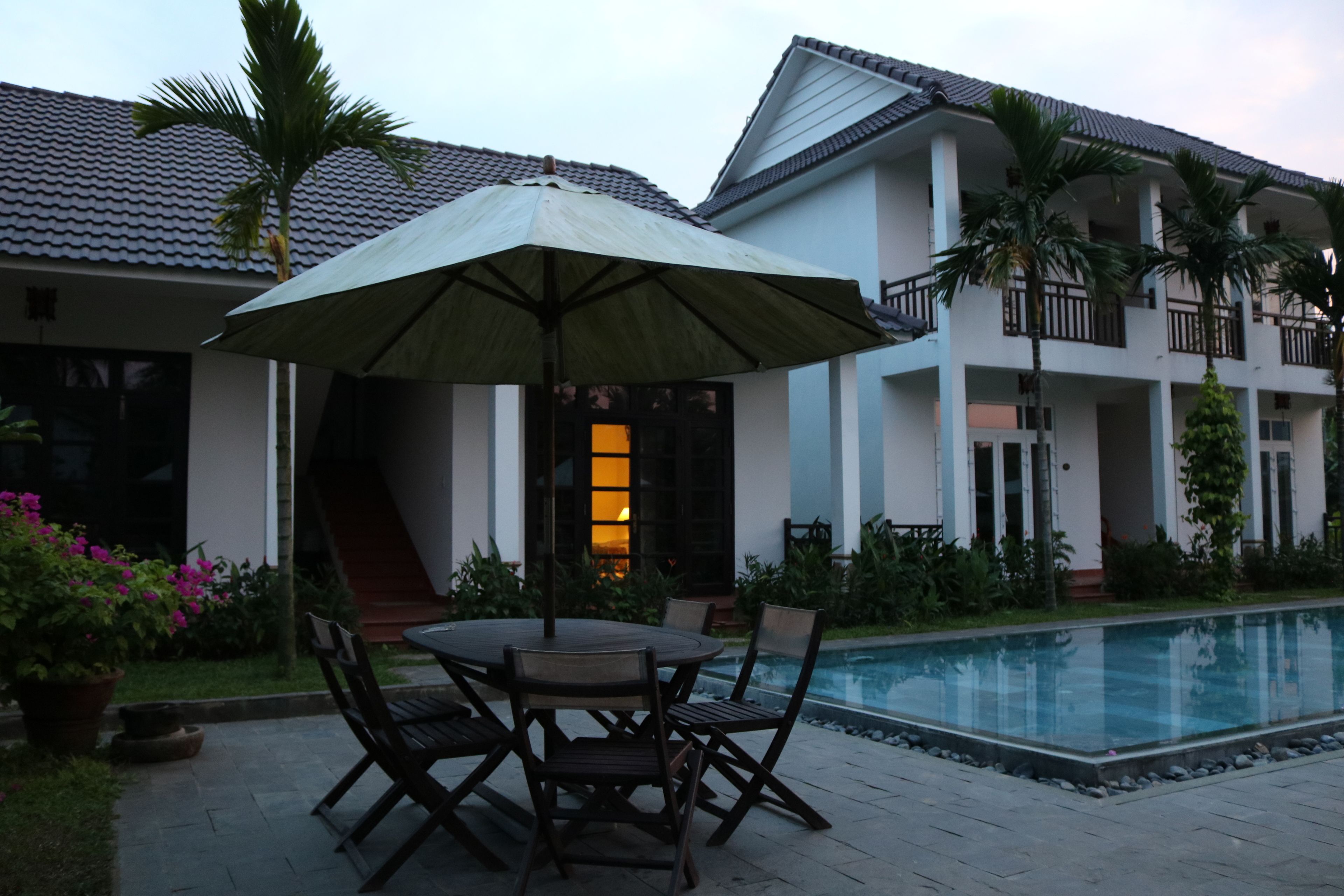 A poolside area with a table and chairs, a large umbrella, and a building with balconies and a swimming pool.