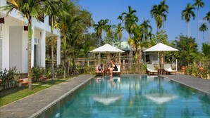 Una piscina con sillones de descanso y sombrillas, rodeada de palmeras y un edificio blanco.