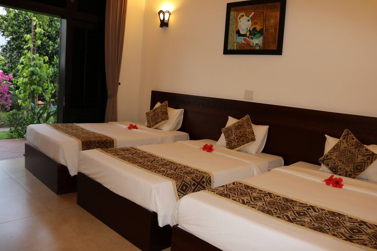 Habitación de hotel con tres camas, cada una con colchas y almohadas estampadas, un cuadro en la pared y vistas a zonas verdes.